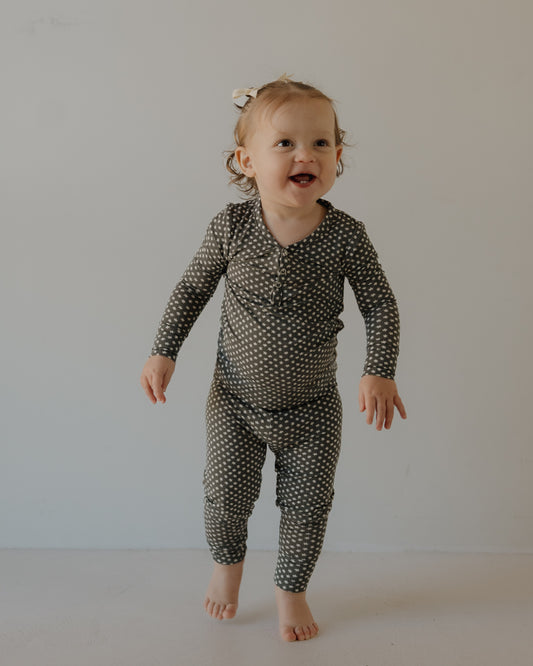A smiling toddler with light brown hair and a white bow wears forever french baby Bamboo Two Piece Pajamas in Green Flowers, standing barefoot against a plain light-colored background.