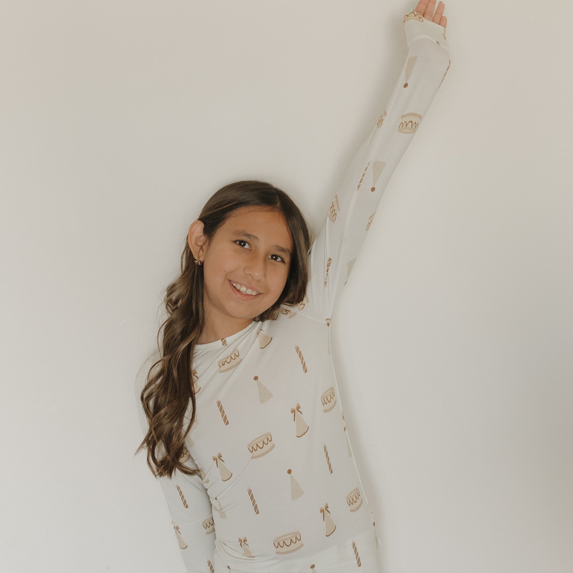 A cheerful girl with long brown hair wears the "Child Sleepy Time Set | Birthday Girl" from forever french baby, featuring whimsical birthday cake and party hat prints on soft cream bamboo fabric, raising one arm against a plain white wall. This cozy set is hypo-allergenic and perfect for sensitive skin, combining both comfort and playful style.