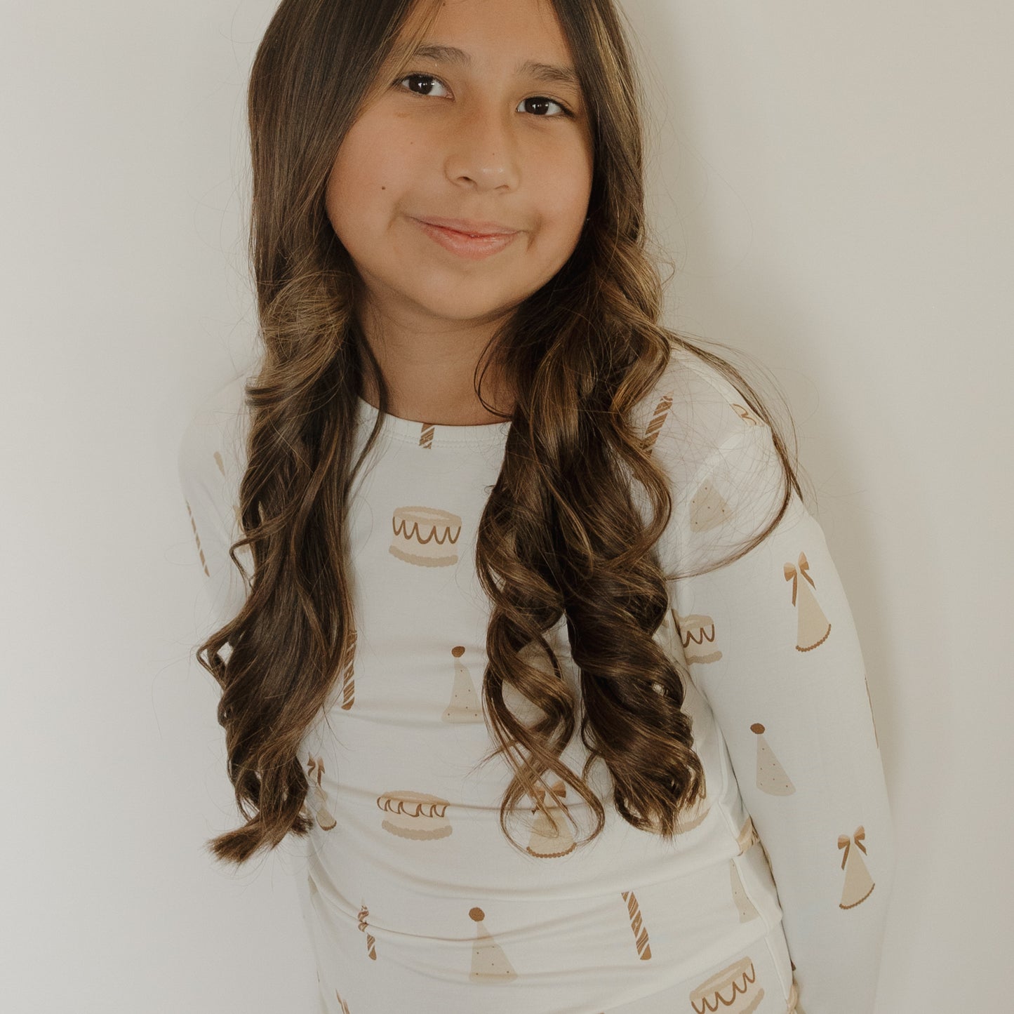 A smiling girl with long, wavy brown hair is wearing the forever french baby Child Sleepy Time Set | Birthday Girl—a soft bamboo set featuring cake, candle, and party hat patterns—as she stands in front of a plain white background.