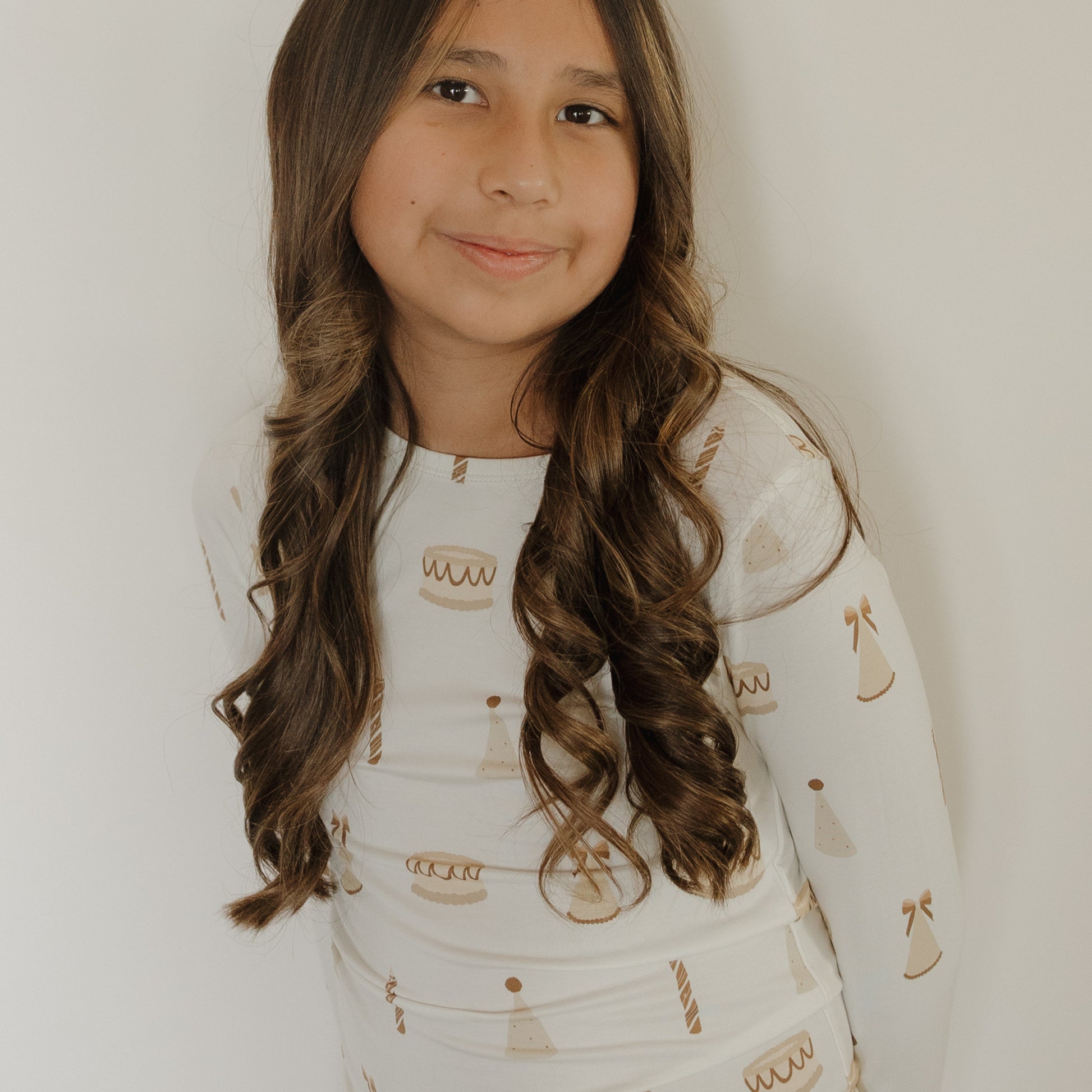 A smiling girl with long, wavy brown hair is wearing the forever french baby Child Sleepy Time Set | Birthday Girl—a soft bamboo set featuring cake, candle, and party hat patterns—as she stands in front of a plain white background.