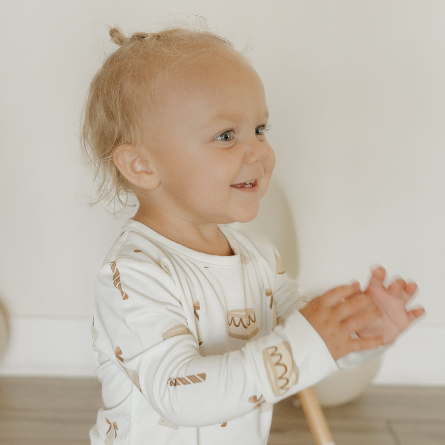 A smiling blond toddler with a top knot wears the forever french baby Child Sleepy Time Set | Birthday Girl, clapping her hands while standing indoors on a wooden floor.