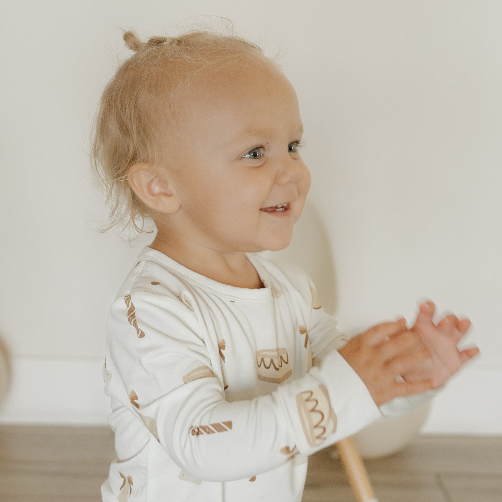 A smiling blond toddler with a top knot wears the forever french baby Child Sleepy Time Set | Birthday Girl, clapping her hands while standing indoors on a wooden floor.