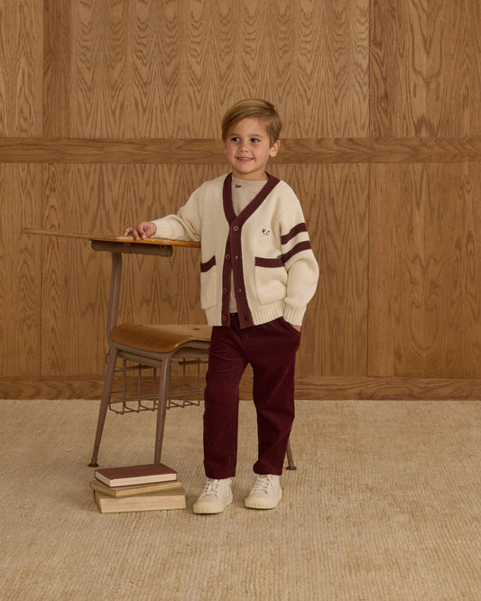 A young child wearing the Rylee + Cru Haywood Cardigan in natural stands beside a small wooden desk with books scattered on the floor. Set against a wood-paneled background, the child smiles while resting one hand on the desk.