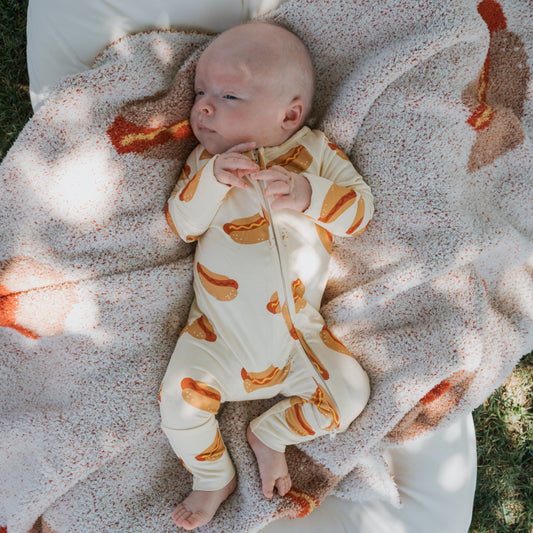 Outdoors, a baby lies on a soft, patterned blanket wearing forever french baby Bamboo Zip Pajamas | Hot Dogs—a cream onesie with hot dog prints—gazing upward with hands touching at the chest.