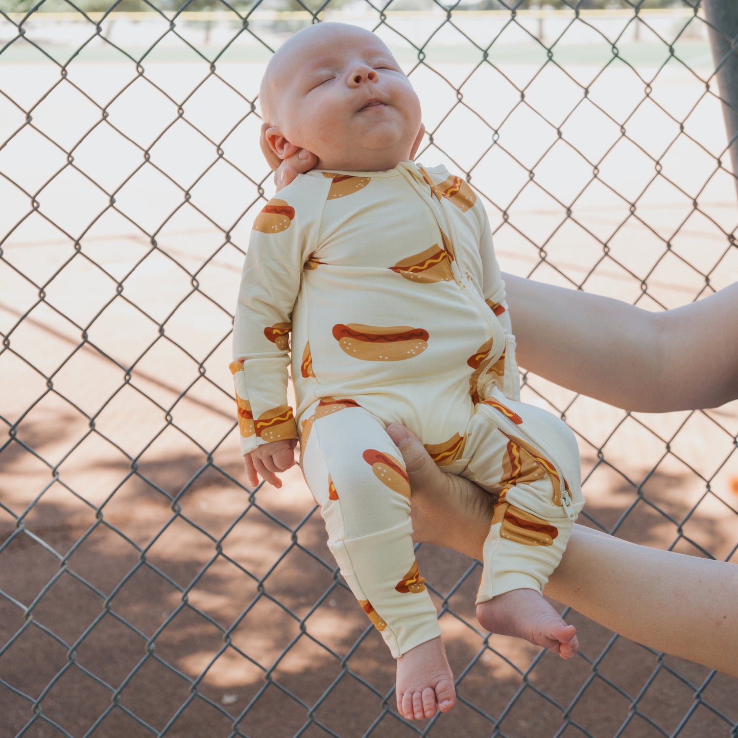 A baby in forever french baby's cream-colored Bamboo Zip Pajamas | Hot Dogs is held up by a chain-link fence at a baseball field, eyes closed and arms relaxed.