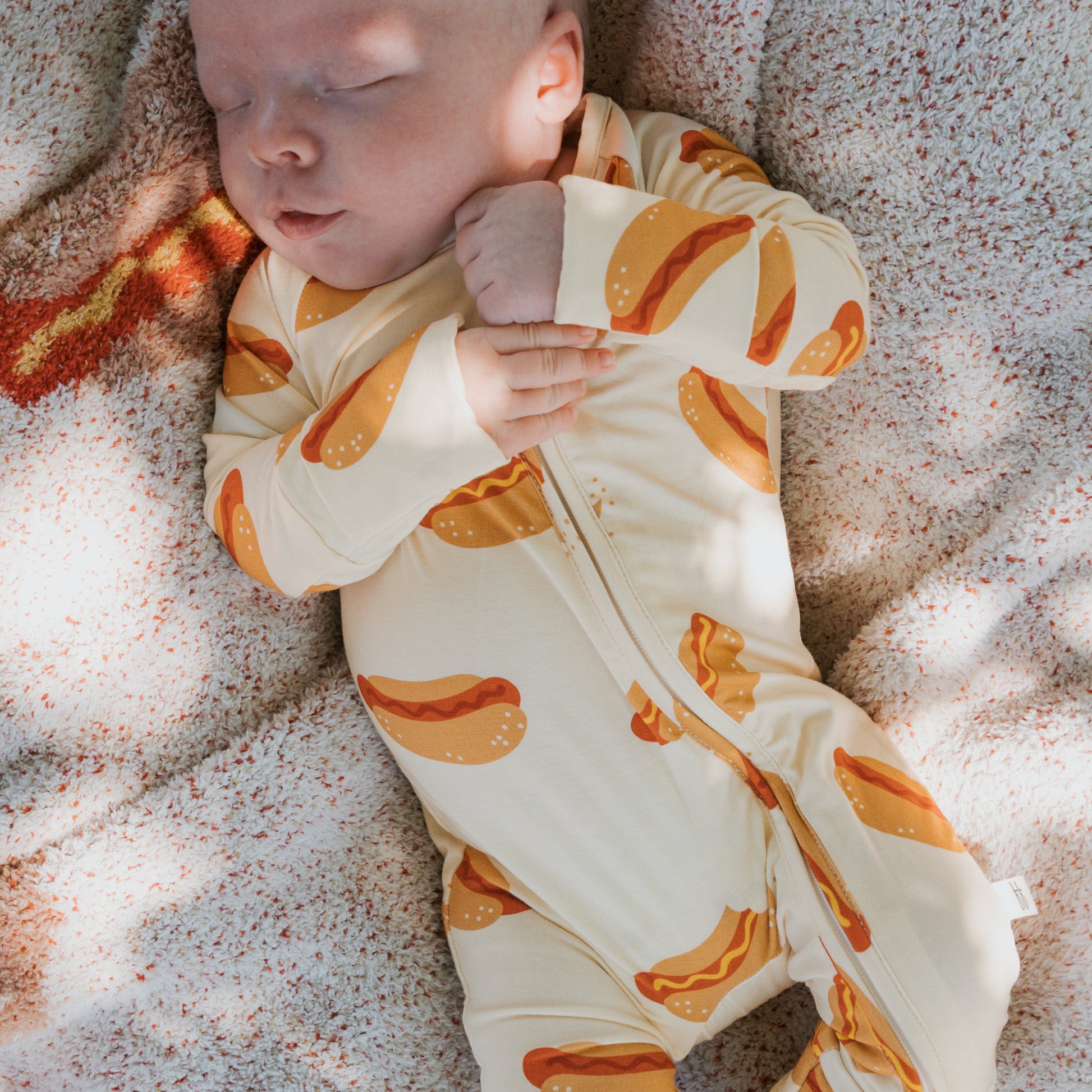 A baby sleeps on a textured blanket, dressed in forever french baby's Bamboo Zip Pajamas | Hot Dogs—hypoallergenic and designed for ultimate comfort.
