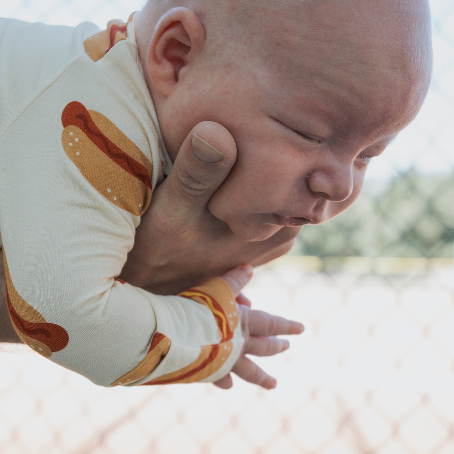 A baby in forever french baby Bamboo Zip Pajamas | Hot Dogs is gently held outdoors by an adult hand, with a blurry fence in the background.