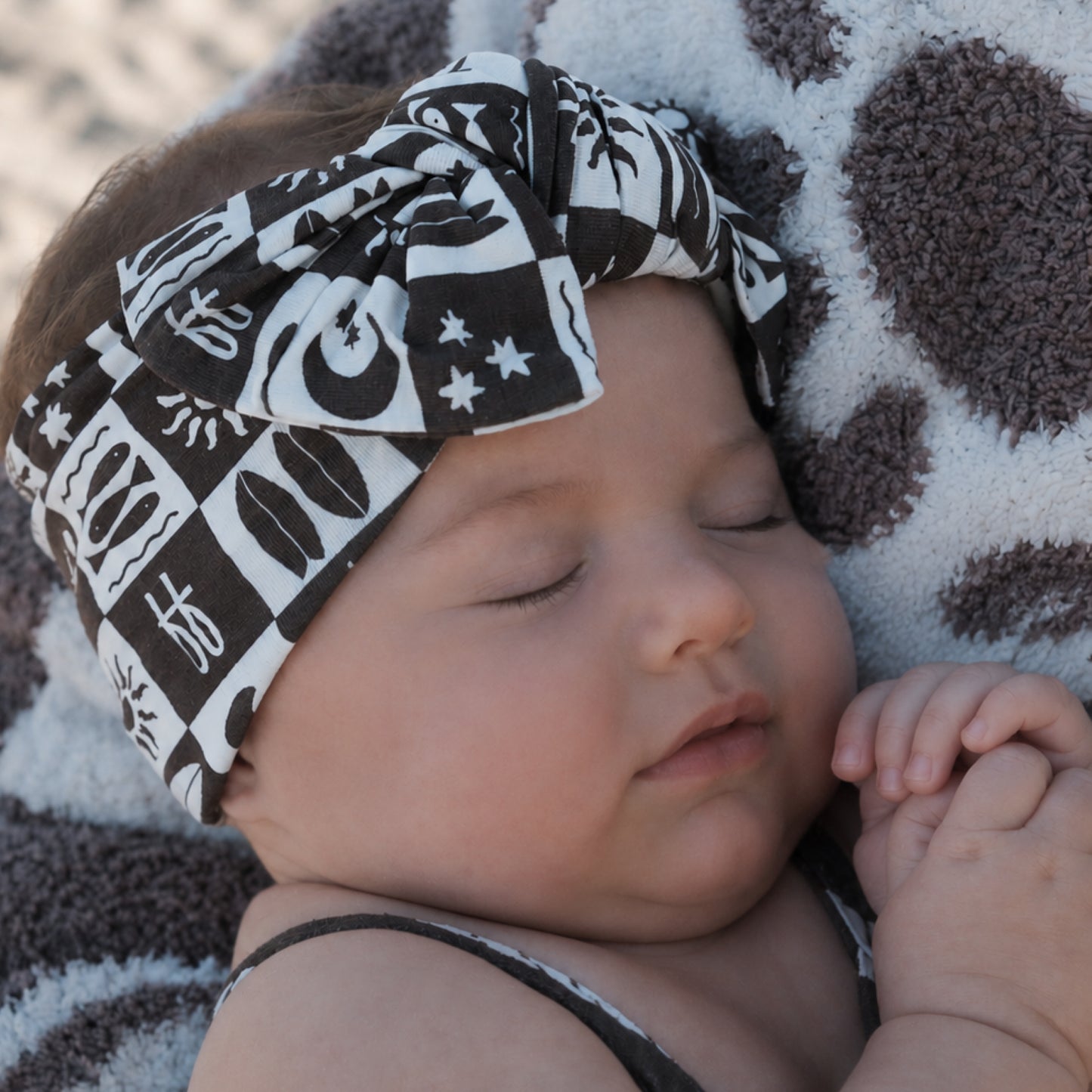 A baby with closed eyes sleeps peacefully, wearing the forever french baby Bamboo Head Wrap | Summer Check, featuring a large bow, and resting against a soft, spotted blanket.