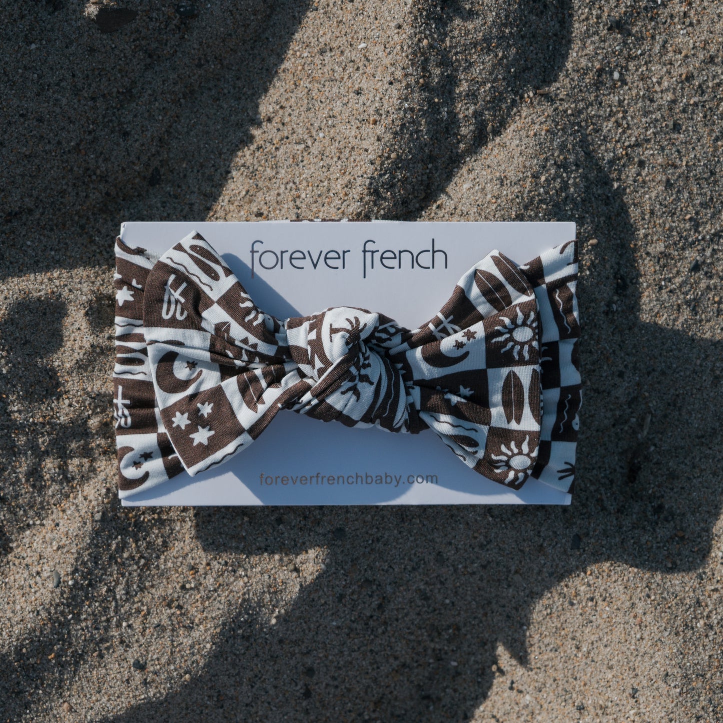 A Bamboo Head Wrap | Summer Check from forever french baby, featuring brown and white patterns with symbols, rests on sandy ground with shadows. The adjustable wrap is displayed on branded packaging.