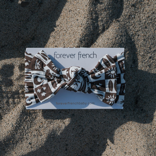 A Bamboo Head Wrap | Summer Check from forever french baby, featuring brown and white patterns with symbols, rests on sandy ground with shadows. The adjustable wrap is displayed on branded packaging.