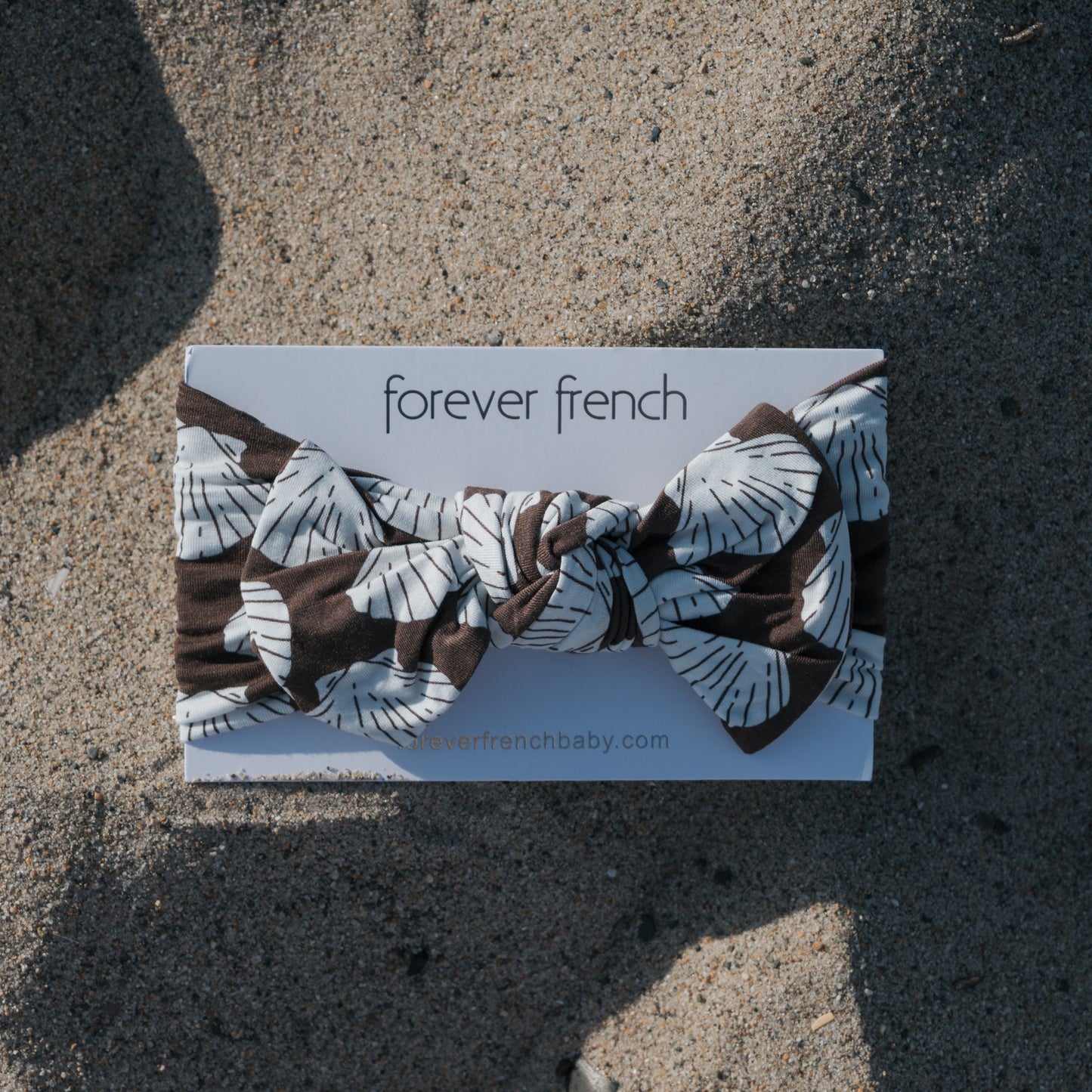 The Bamboo Head Wrap | Seashells from forever french baby, featuring a brown and white leafy pattern, is displayed on a branded card placed on sandy ground.