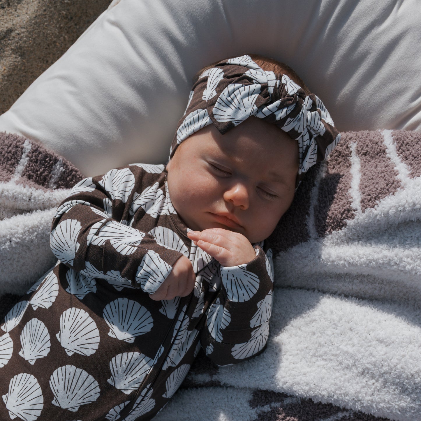 A baby in forever french baby Bamboo Zip Pajamas | Seashells, featuring white seashell patterns and a matching headband, sleeps on a white pillow wrapped in a striped blanket.
