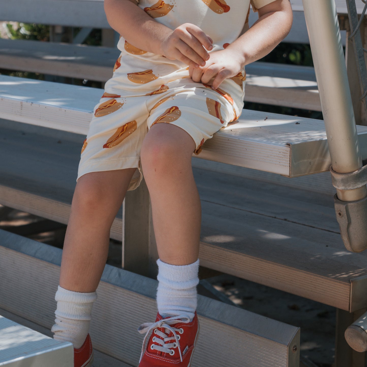 A child sits on bleachers wearing the forever french baby Bamboo Short Set | Hot Dogs—breathable bamboo shirt and shorts for all-day comfort. The child's face is not visible, paired with white socks and red sneakers.