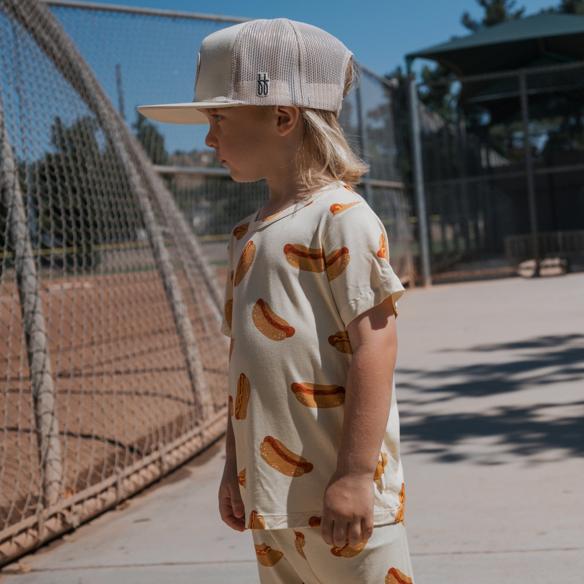 A young child in the forever french baby Bamboo Short Set | Hot Dogs and a beige baseball cap stands by a chain-link fence on a sunny day at the baseball field.