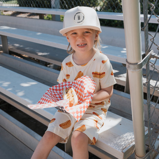A smiling young child sits on bleachers, wearing the forever french baby Bamboo Short Set | Hot Dogs. They hold a hot dog in a checkered tray, sunlight filters in, and a chain-link fence is visible behind them.