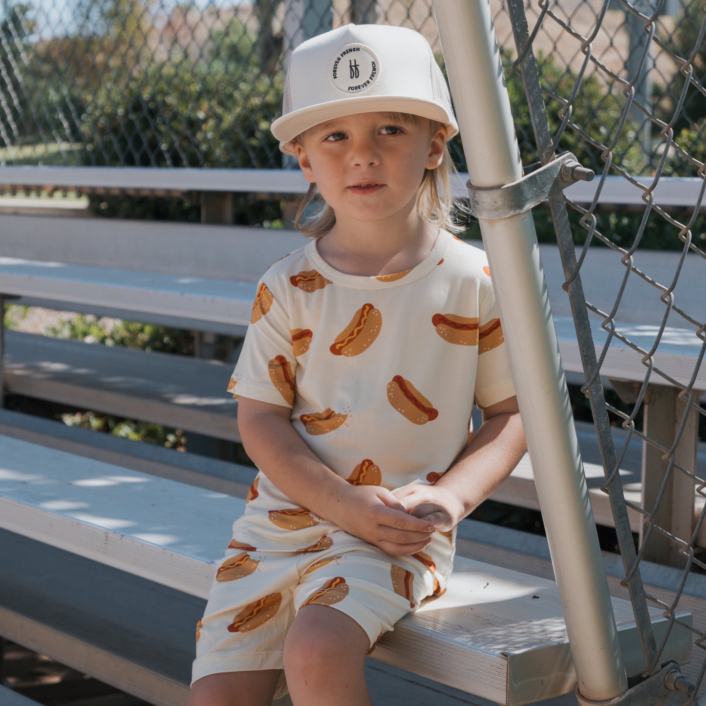 A young child in the forever french baby Bamboo Short Set | Hot Dogs and a beige cap sits on silver bleachers by a chain-link fence, enjoying a sunny day outdoors.