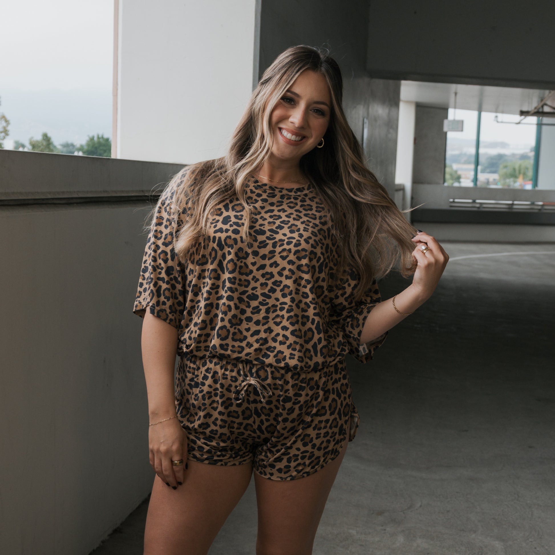 A woman with long brown hair smiles and stands in a parking garage wearing the forever french baby Women's Bamboo Short Set in Leopard, holding a strand of her hair with one hand.