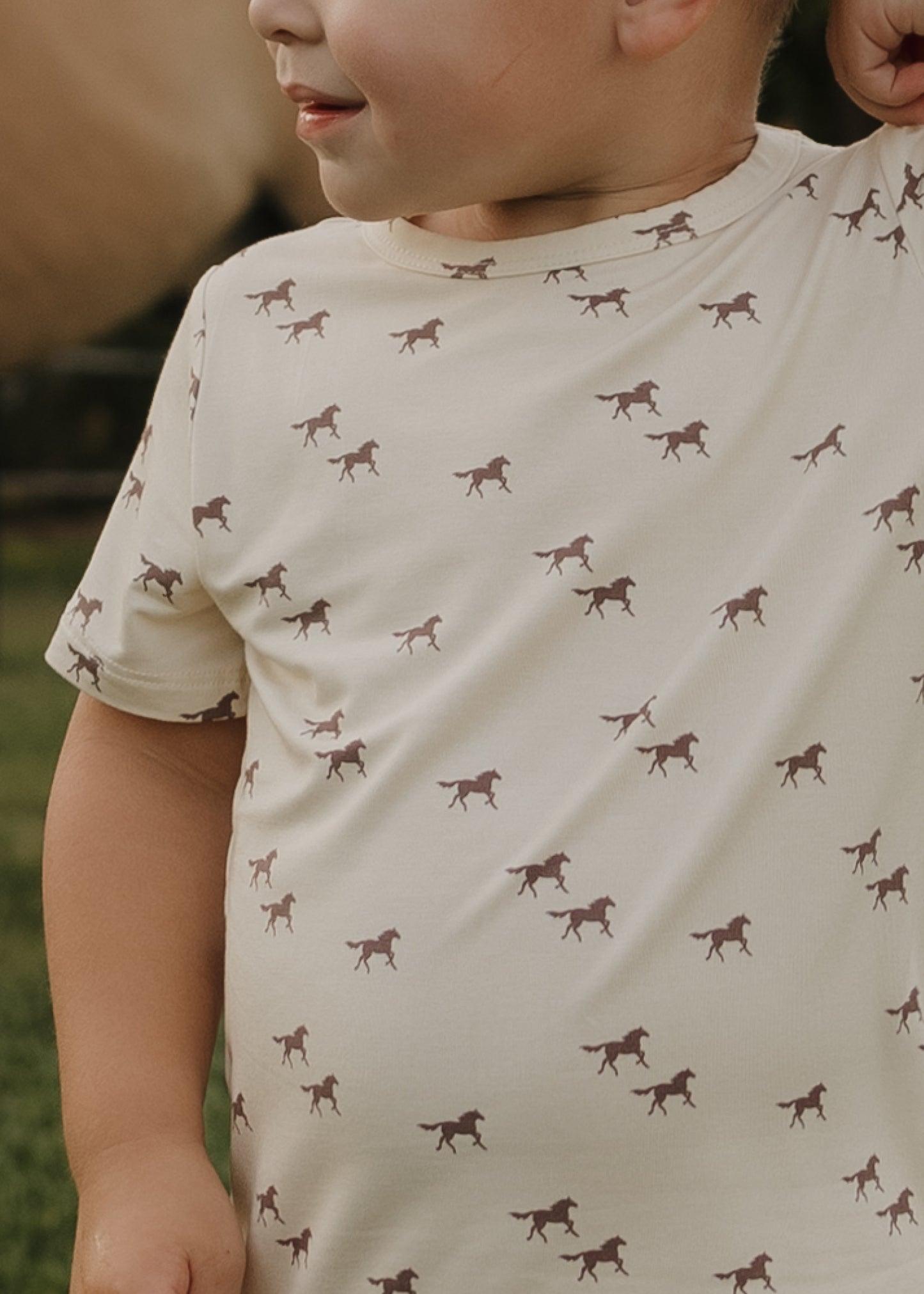 A young child wears the forever french baby Bamboo Short Set | Wild Horses, a breathable kids' outfit with a brown horse pattern. Only their upper body and part of their face are visible, with blurred greenery in the background.