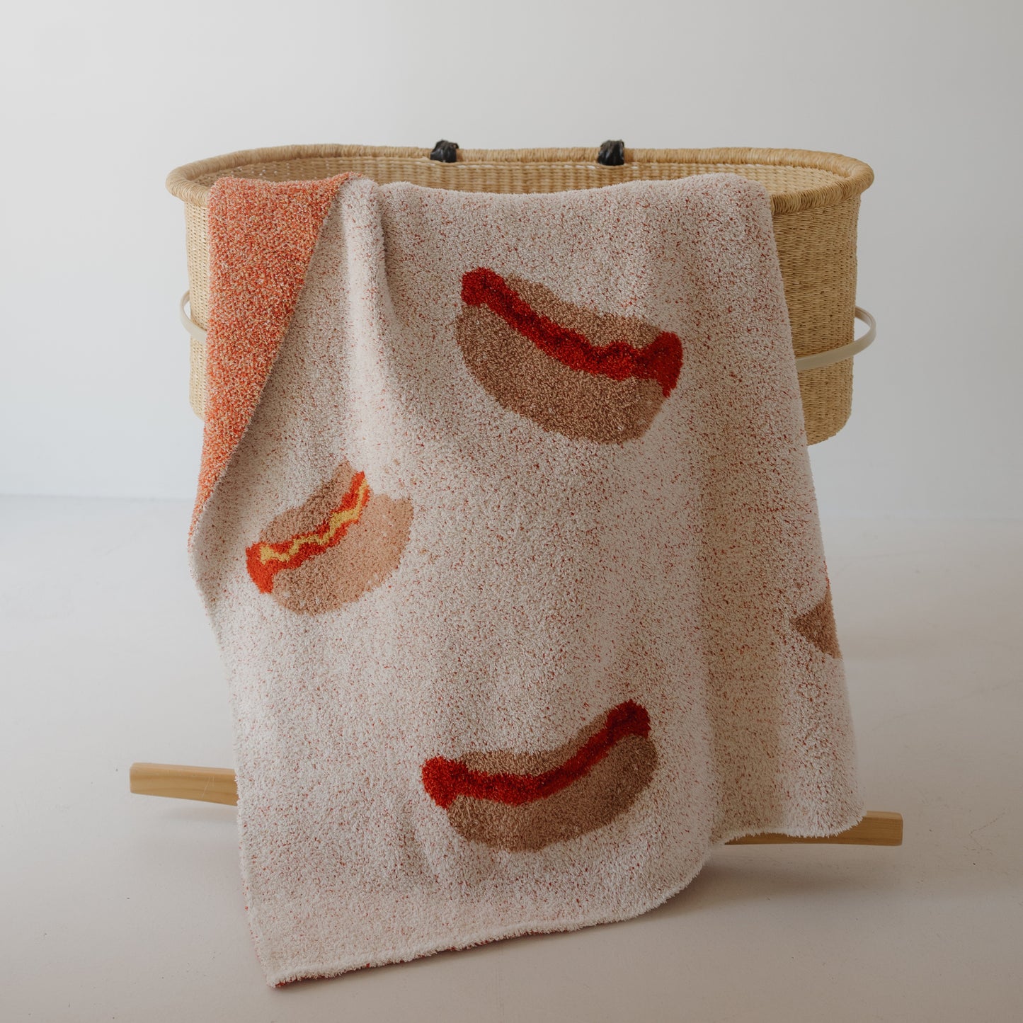 The forever french baby Plush Blanket | Hot Dogs—a beige knit microfiber, Oeko-Tex certified—features playful hot dog designs and is shown draped over a woven basket against a white background.