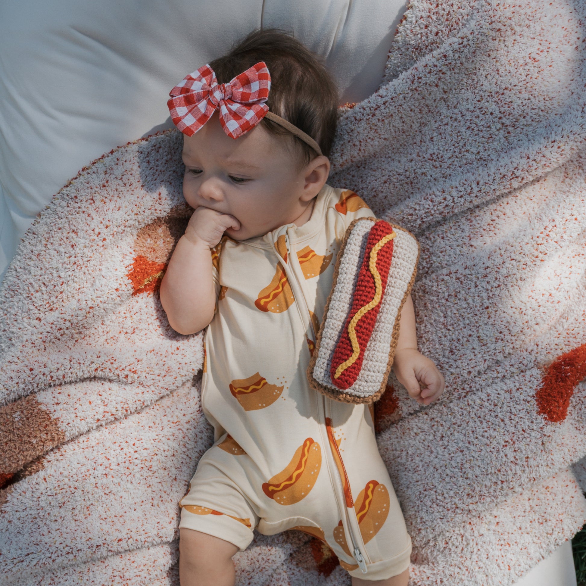 A baby in a hot dog print romper and red plaid bow headband lies on a blanket, sucking their hand and holding the *SHIPS IN 1 WEEK* Knit Rattle | Hot Dog from forever french baby.