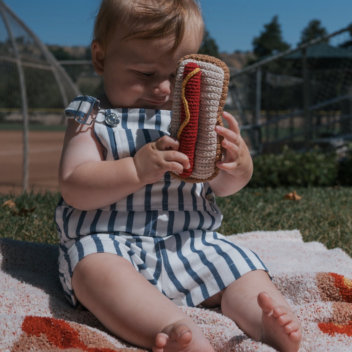 A baby in a blue and white striped romper sits on a blanket outdoors, holding the forever french baby *SHIPS IN 1 WEEK* Knit Rattle | Hot Dog—an adorable summer toy.