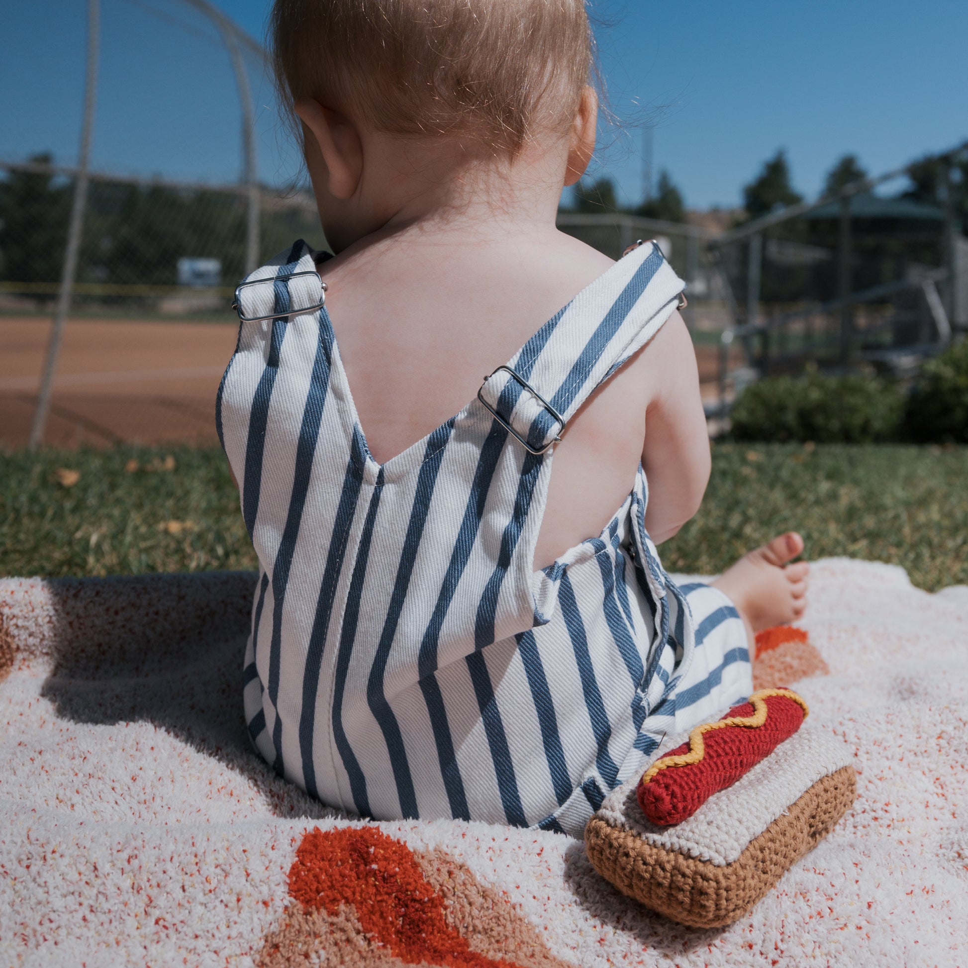 A baby in a blue and white striped romper sits on a blanket at a sunny baseball field, with a forever french baby *SHIPS IN 1 WEEK* Knit Rattle | Hot Dog beside them.