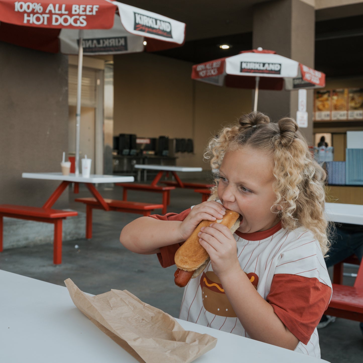 A young child with curly blonde hair, wearing a forever french baby Child Cotton Tee | Hot Dogs, happily eats a large hot dog at an outdoor table in front of a Kirkland food stand with red umbrellas and hot dog signs.