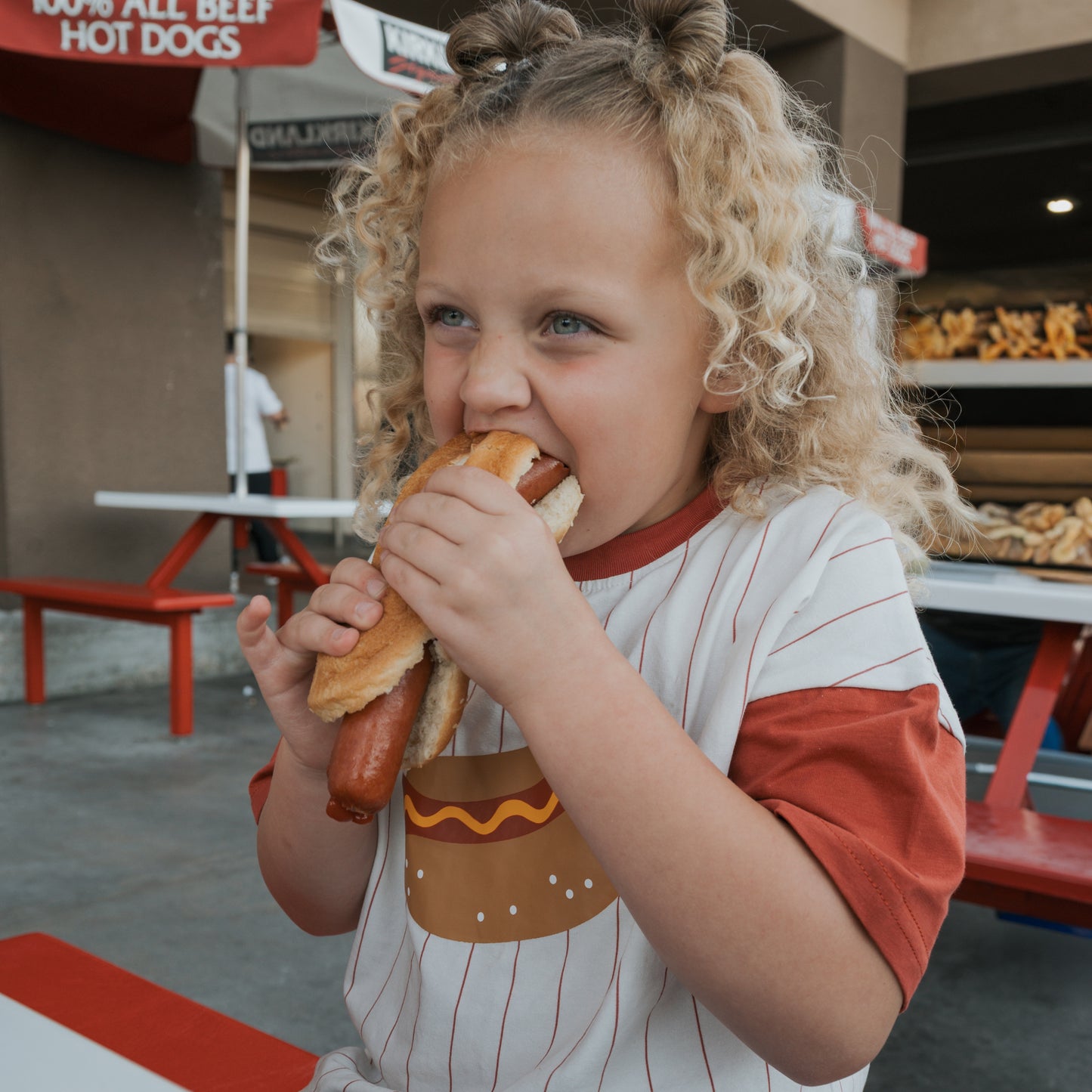 A young child in a forever french baby Child Cotton Tee | Hot Dogs takes a big bite of a hot dog while sitting at a red and white picnic table outside a food stand.
