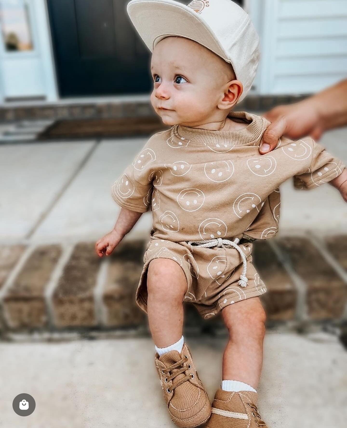 A small child, dressed in a Forever French Baby Just Smile Tan short set with smiley face patterns, beige sneakers, and a beige cap, sits on a pavement in front of a building. An adult's hand gently steadies the child from the side.