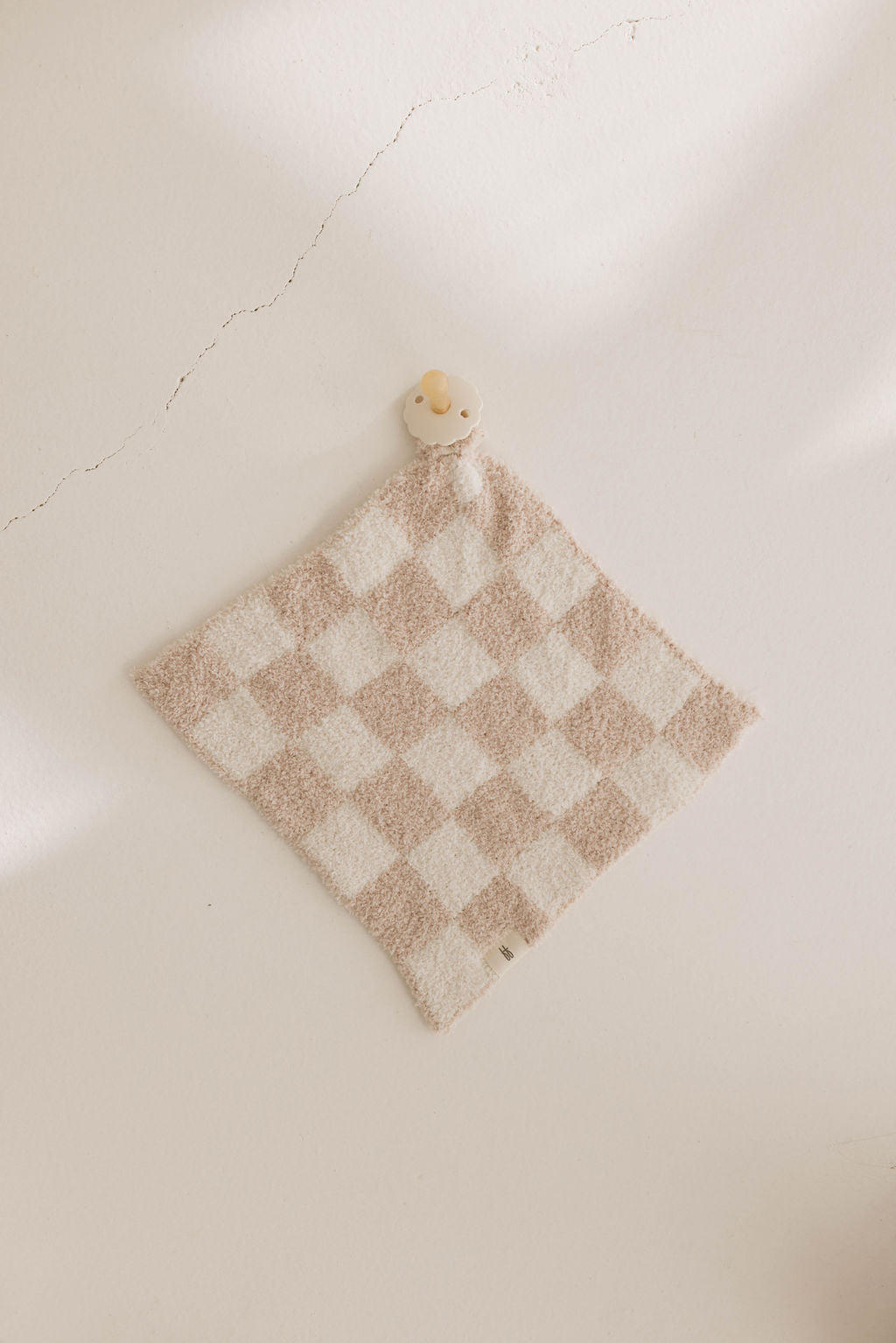 A soft taupe and white checkerboard pattern cloth from forever french baby, named Lovey, is laid flat on a surface with a small stuffed animal resembling a bear placed at the top corner. The wall in the background has a visible crack. The lighting is soft and gentle, casting mild shadows.