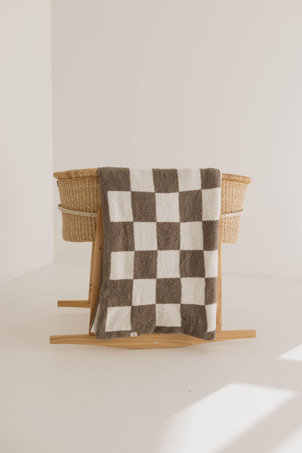 A wooden rocking chair holds a Plush Blanket | Mushroom Checkerboard by forever french baby, made of Oeko-Tex certified materials. The wicker-style chair is placed against a plain, light-colored backdrop. Soft daylight filters in, creating a serene and minimalist scene.