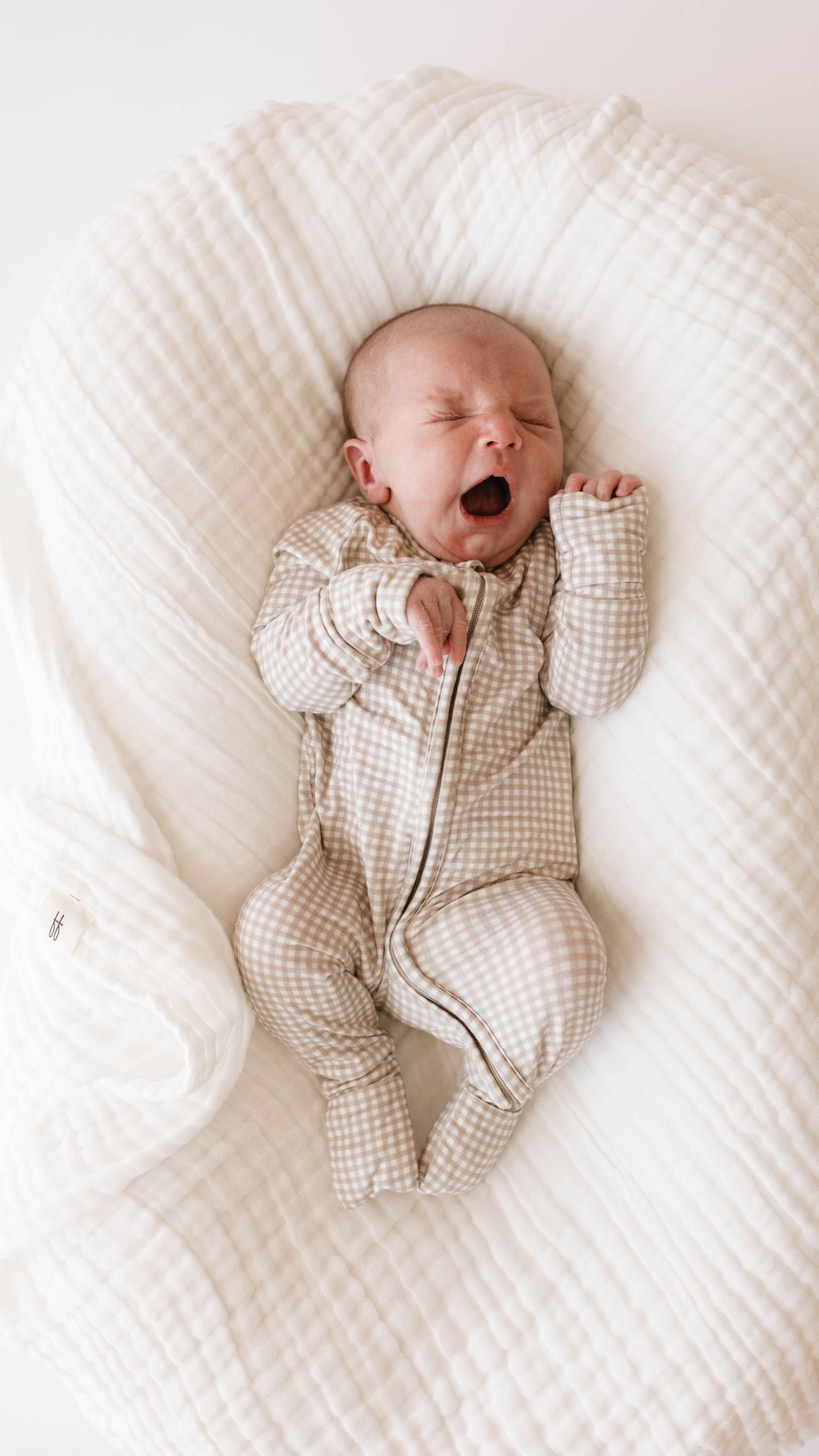 A baby lies on a soft white crib sheet from forever french baby, with eyes closed and mouth open, appearing to yawn or cry. Wearing a beige and white checkered onesie, the baby's hands are positioned with one clenched into a fist and the other slightly raised.