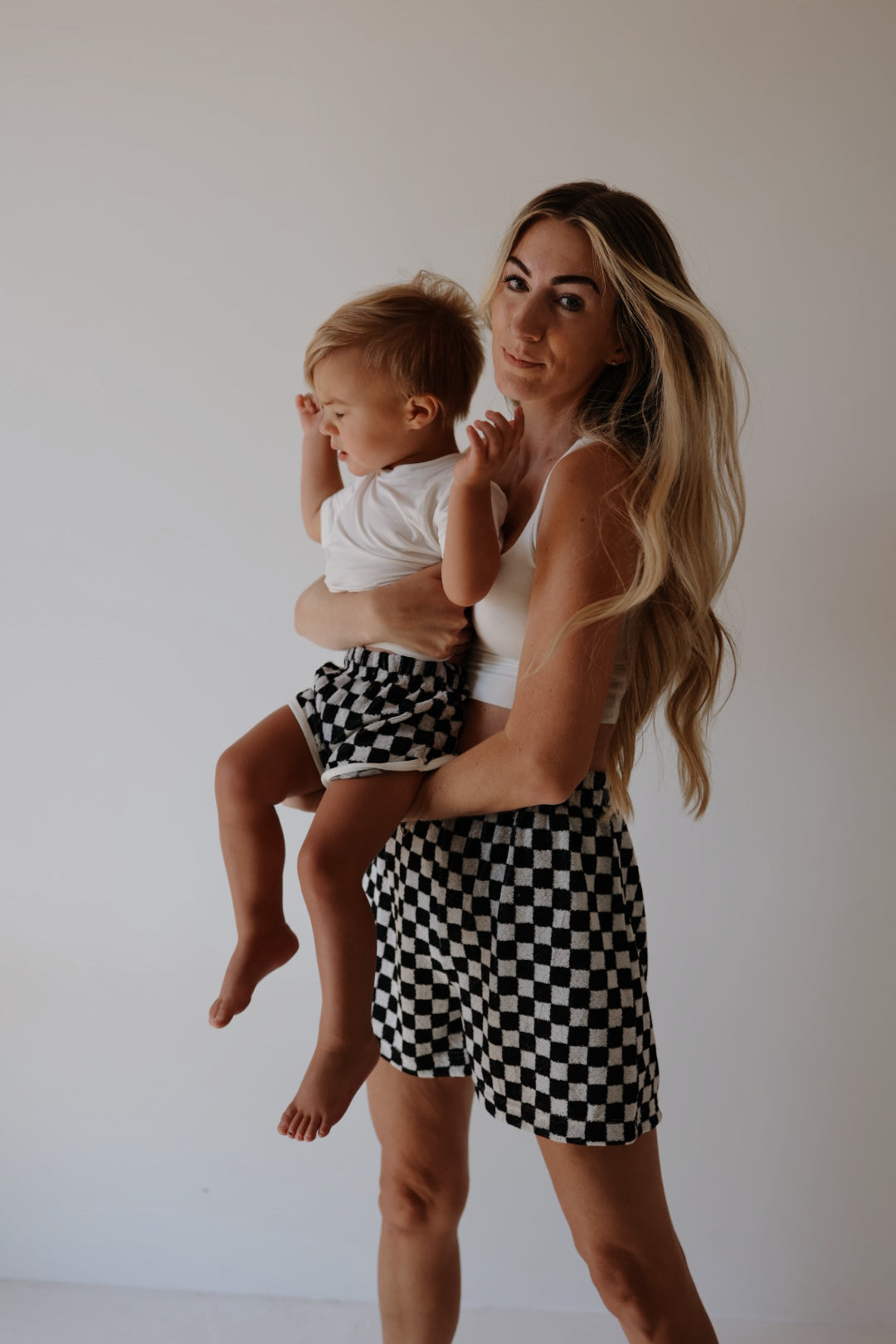 A long-haired woman dressed in a summer wardrobe featuring a white top and forever french baby's Women's Terry Shorts in Black + White Checkerboard holds a toddler wearing a white shirt and matching checkered shorts. The background is plain white, creating a clean and minimalistic look.