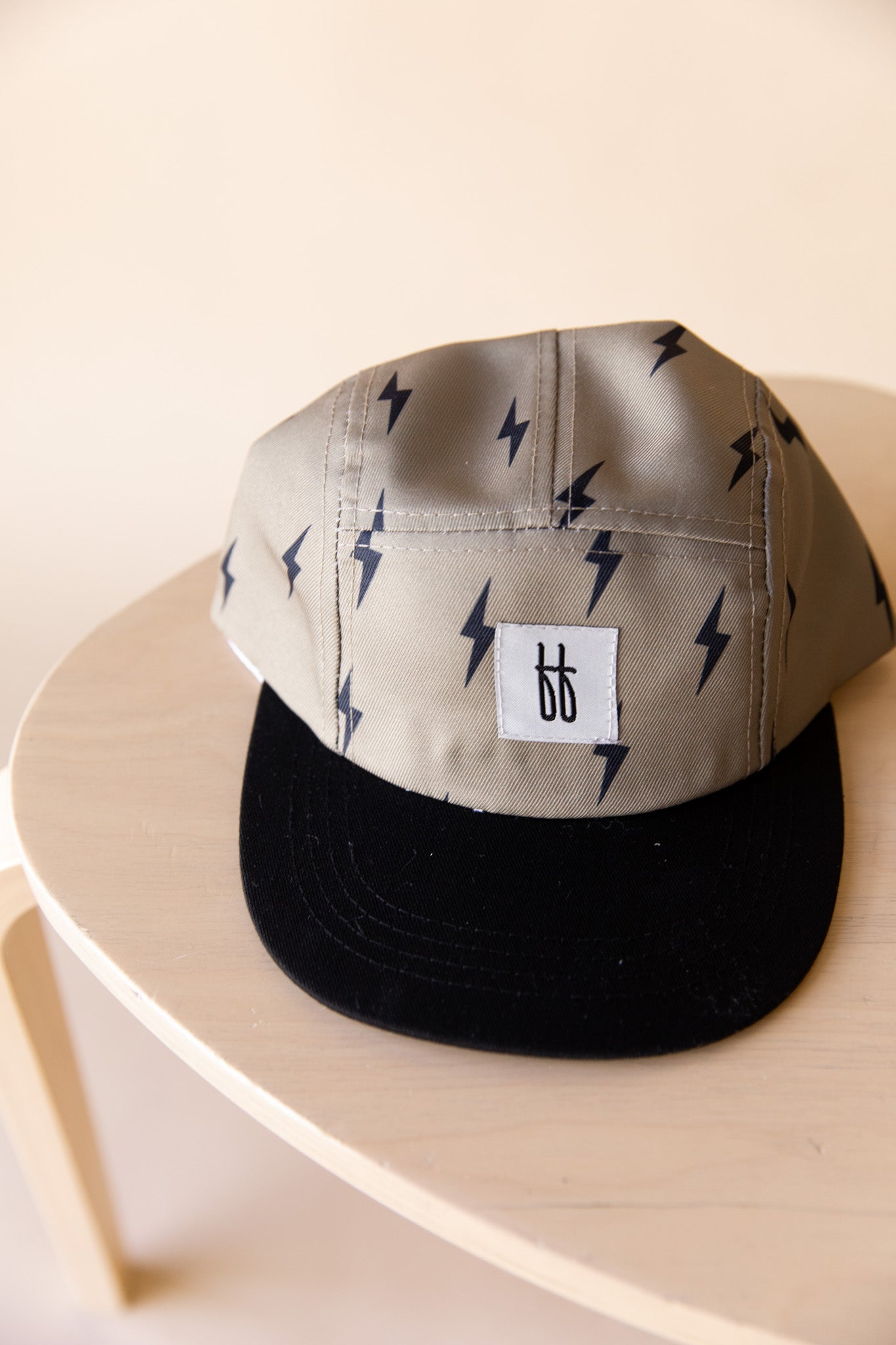 A beige Snap Back | Lightning Bolt baseball cap from forever french baby, featuring black lightning bolt patterns and a black brim, rests on a light wooden table. The front of the cap sports a white square patch with the initials "bb" in black text.