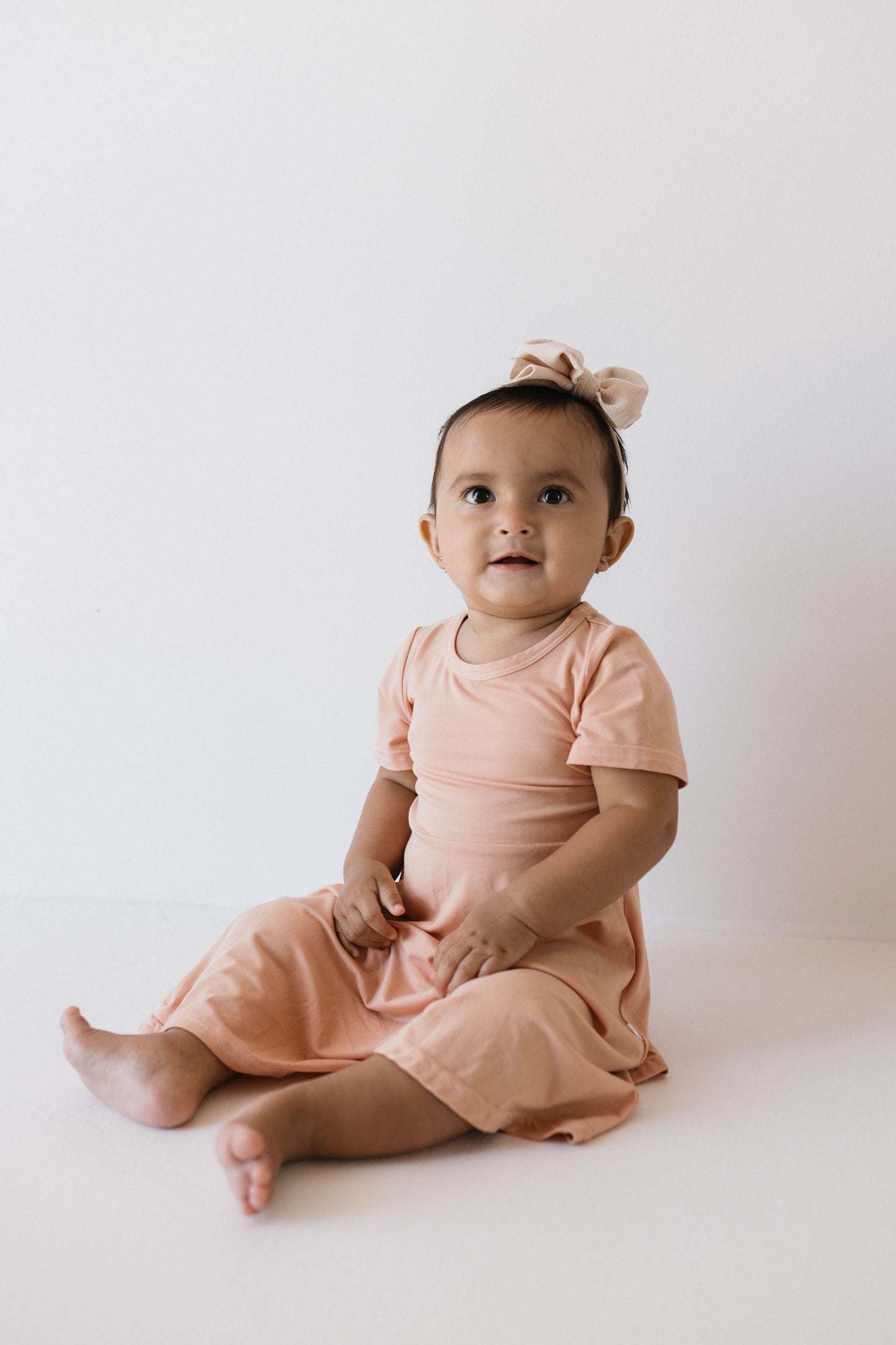 A baby wearing a "Bamboo Dress | Clay" from forever french baby, crafted from breathable fabric and paired with a matching bow, sits on the floor against a plain white background. The baby looks slightly upward with a curious expression.