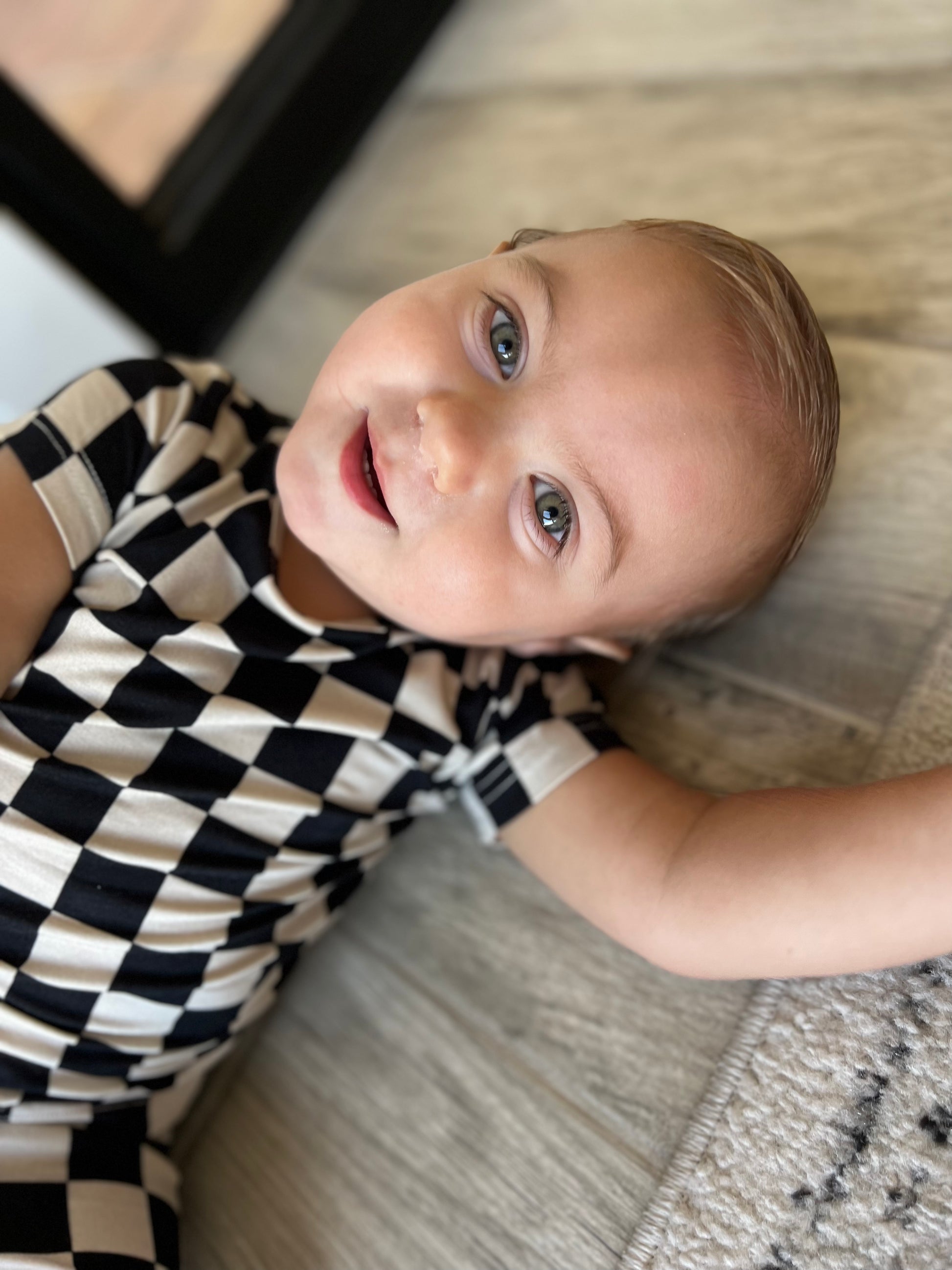 A baby with light eyes and fair skin is lying on a patterned surface. Wearing the breathable Bamboo Short Set in Black Checkerboard by forever french baby, the baby smiles with an arm outstretched. The background includes a wooden floor and part of a carpet, enhancing the cozy ambiance.