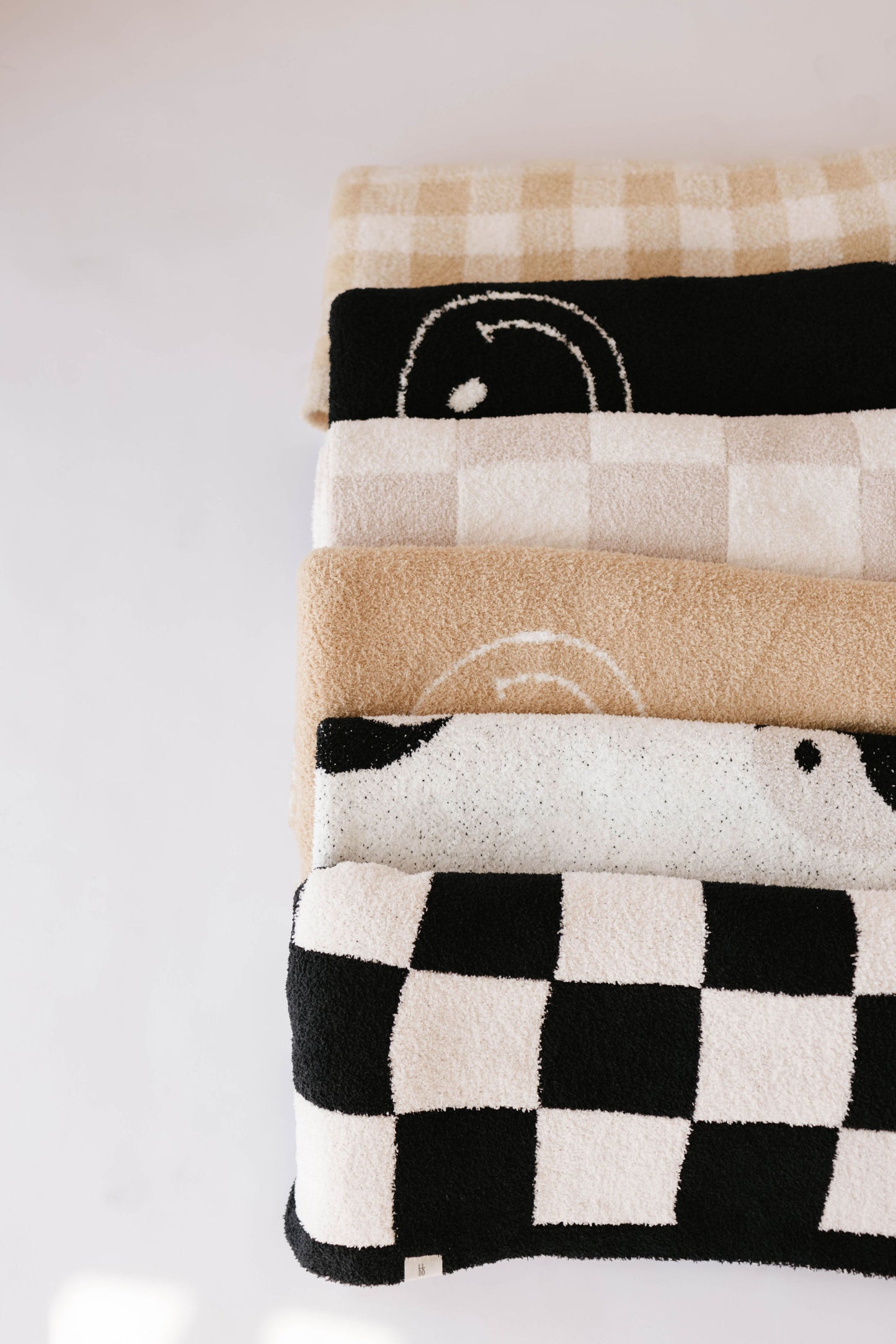 A set of four folded blankets, each showcasing unique checkered patterns and crafted from luxuriously soft microfiber feather yarn. The top blanket boasts a light beige and ivory checkerboard pattern. The second one, directly underneath, is adorned with a smiling face. The bottom two blankets feature beige and black, as well as the Plush Blanket | Black & White Yin Yang design by forever french baby.