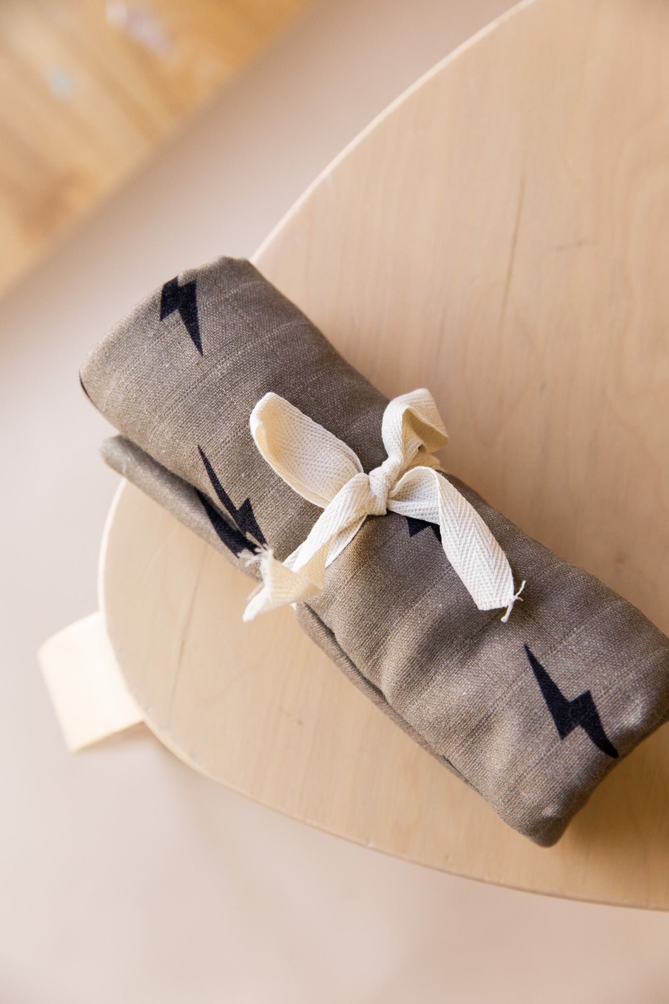 The rolled-up Muslin Swaddle | Lightning Bolt from forever french baby, featuring small black lightning bolt patterns on light brown muslin cotton, is tied with a cream-colored fabric ribbon. The swaddle is placed on a light wooden surface, likely a table or countertop.