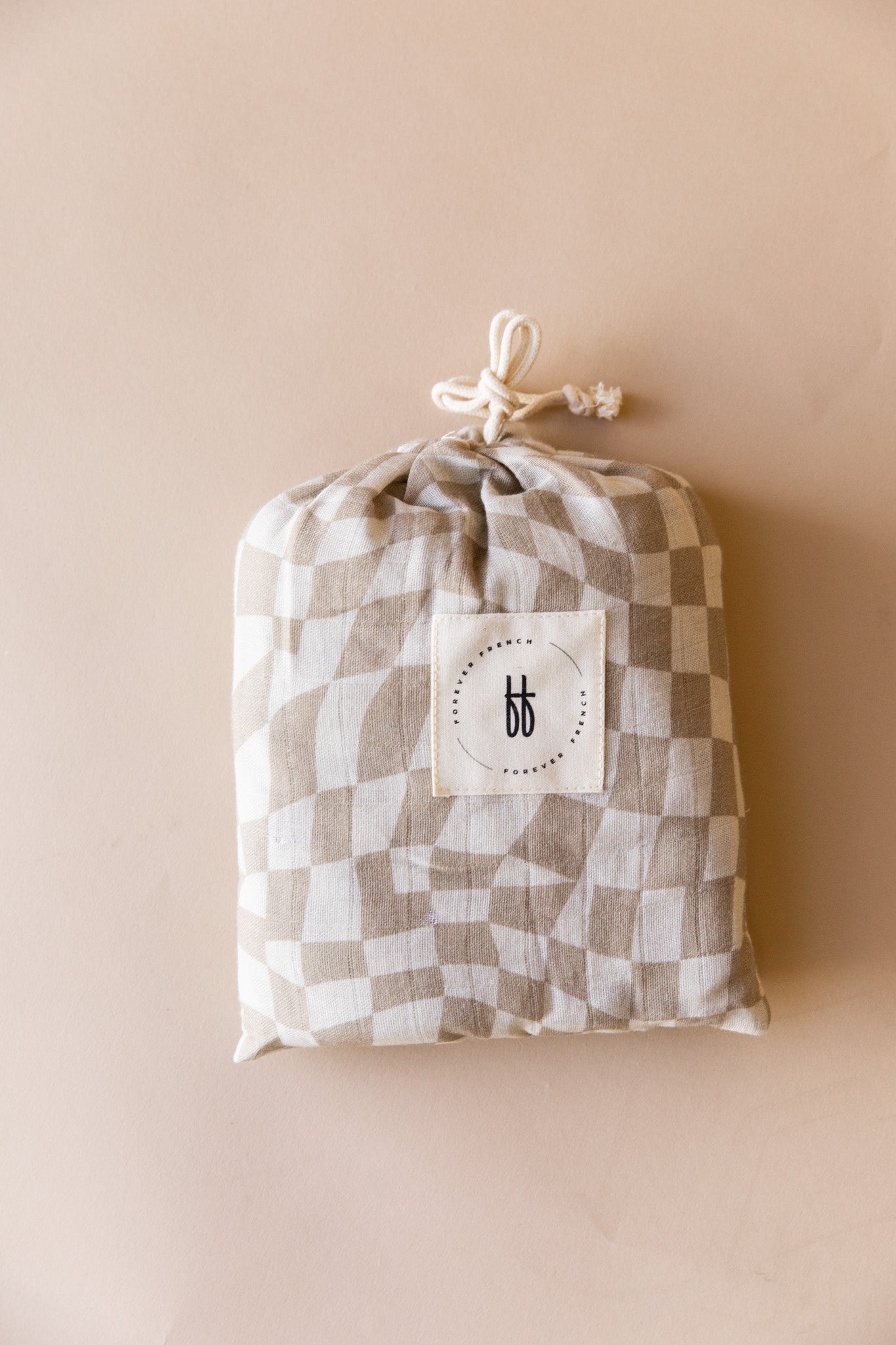 A soft, folded muslin cotton pouch featuring the Wavy Checker pattern in beige and white hues is tied at the top with a drawstring. A small, white rectangular label with a minimalist black logo displaying the initials "bb" from forever french baby is sewn onto the center. The pouch, containing a Muslin Crib Sheet in Wavy Checker, rests on a flat, neutral-colored surface.