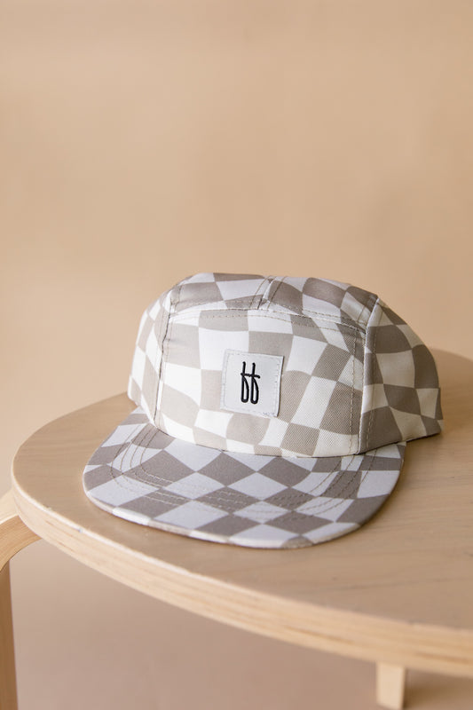 A Forever French Baby "Children's Snap | Wavy Checker" cap, featuring a beige and white checkered design with a white rectangular patch displaying stylized black letters "bb" on the front panel. Designed for ages 4 months to 6 years, the cap rests on a light wooden surface against a beige background. Spot clean only.