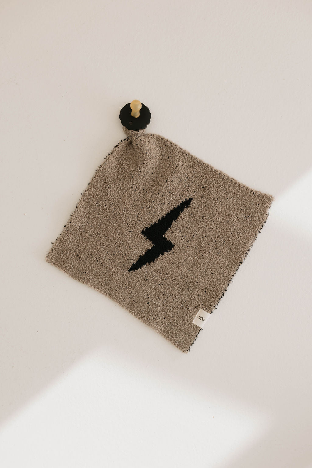 A square cloth featuring a black lightning bolt design in the center is hanging on a white wall. It is attached by a loop to a wooden peg, creating a minimalist display. This Lovey | Lightning Bolt piece from forever french baby appears to be made from the softest lovey fabric, enhancing its gentle charm.