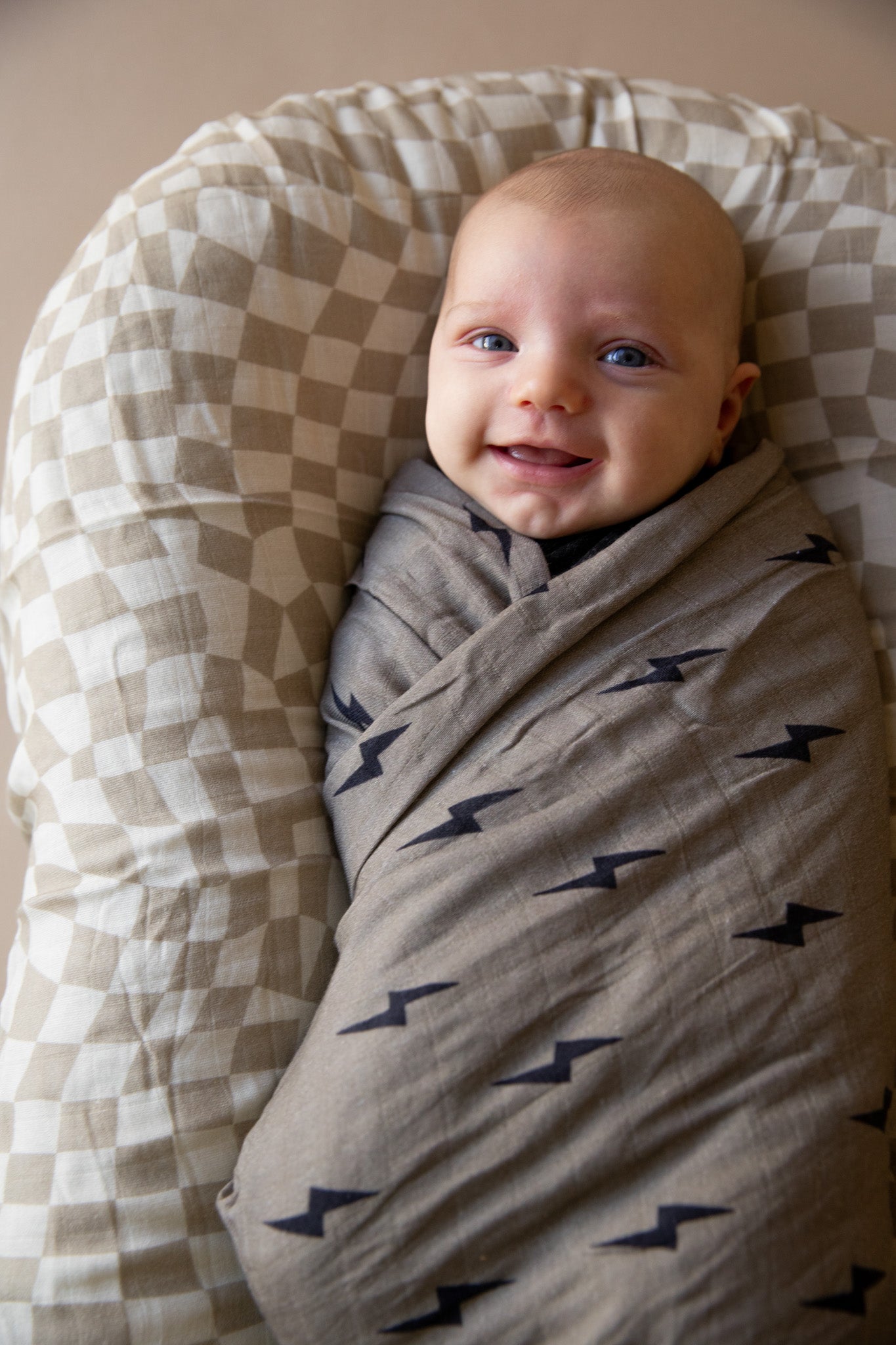 A baby swaddled in a Muslin Swaddle | Lightning Bolt from forever french baby is lying on a patterned cushion. With blue eyes and a smile aimed at the camera, the baby looks cozy and content in their Forever French Baby blanket.