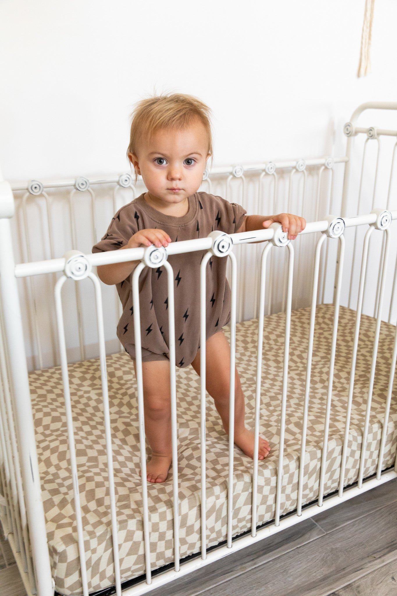 A toddler with short, light brown hair stands in a white metal crib, holding onto the bars. The child is wearing a brown onesie with small black patterns. The crib features a beige and white wavy checkerboard muslin cotton mattress from the "Muslin Crib Sheet | Wavy Checker" by forever french baby. The background showcases a white wall.