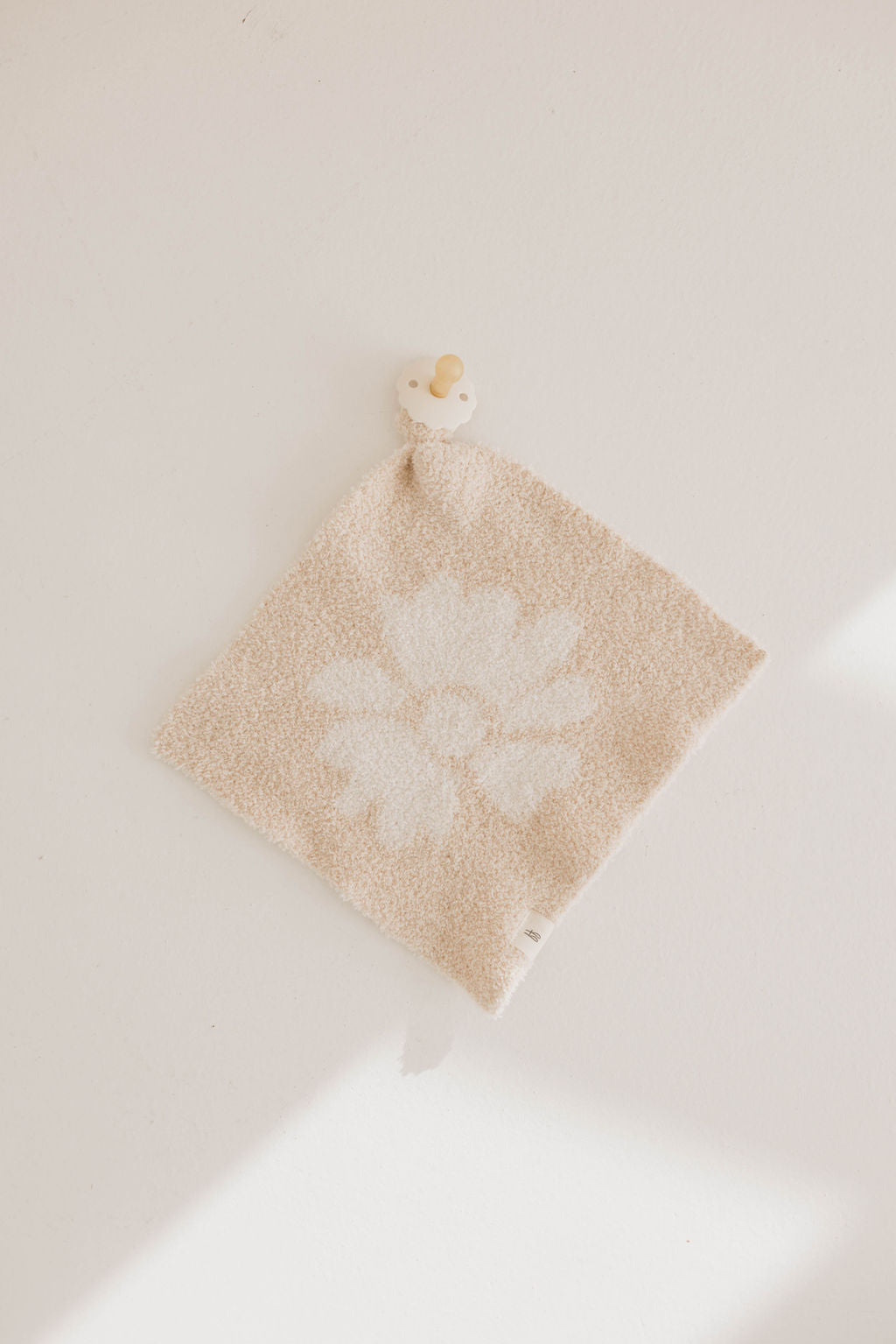 The Lazy Daisy | Lovey by forever french baby is a beige textured microfiber baby lovey with a white flower design, lying flat with a small cream and tan pacifier attached to one corner of the soft fabric.