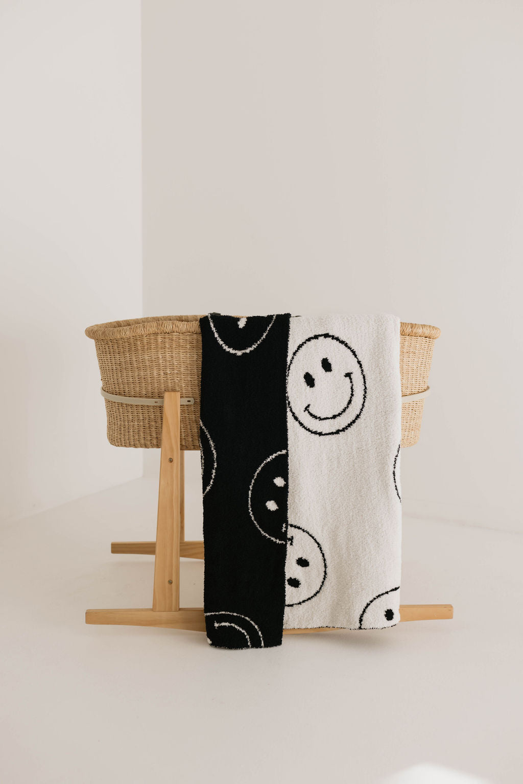 A wooden baby bassinet stands against a plain white wall. Draped over the side is the Plush Blanket | Just Smile Black & White from forever french baby, featuring a dreamy soft split design — half black and half white — each side adorned with smiling face patterns, crafted from luxurious microfiber feather yarn.