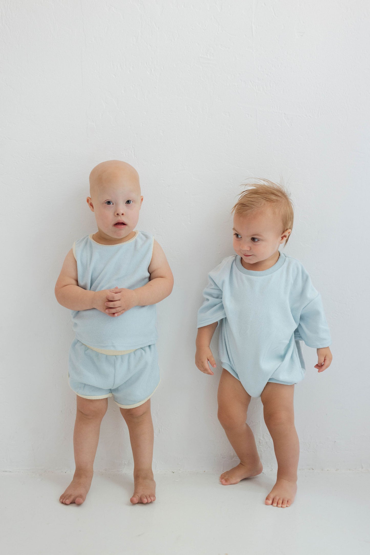 Two toddlers stand against a white wall in light blue outfits. The child on the left wears the "Blue | Terry Tank Set" by forever french baby, while the one on the right sports a long-sleeved shirt. Both are barefoot, gazing in different directions, capturing Forever French Baby charm.