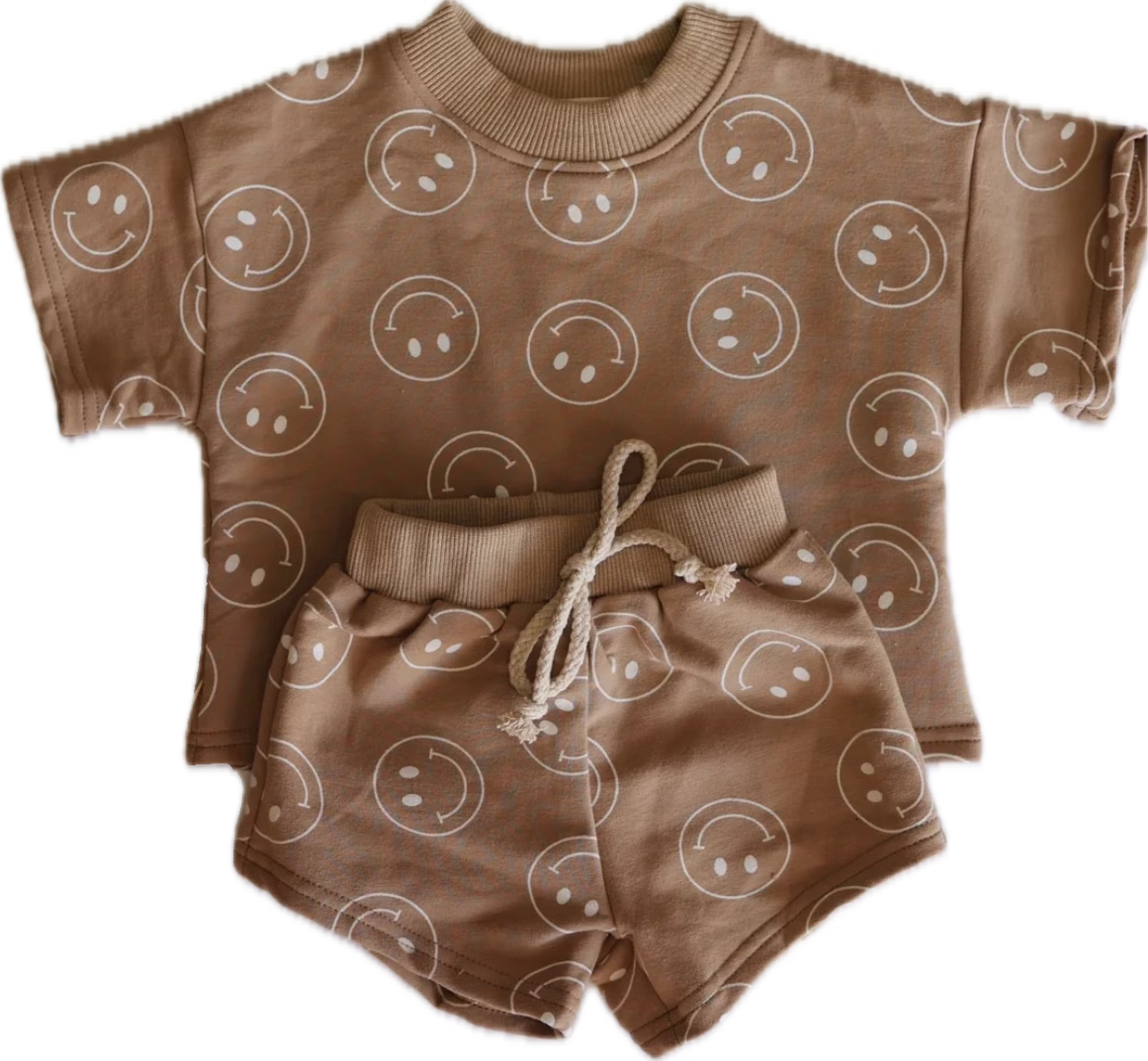 The "Short Set | Just Smile Tan" from forever french baby is a matching beige toddler outfit, featuring a short-sleeve shirt and shorts adorned with an all-over print of white smiley faces. The tan set includes a beige ribbed neckline on the shirt and a drawstring waist on the shorts, making it a perfect addition to any children's wardrobe.