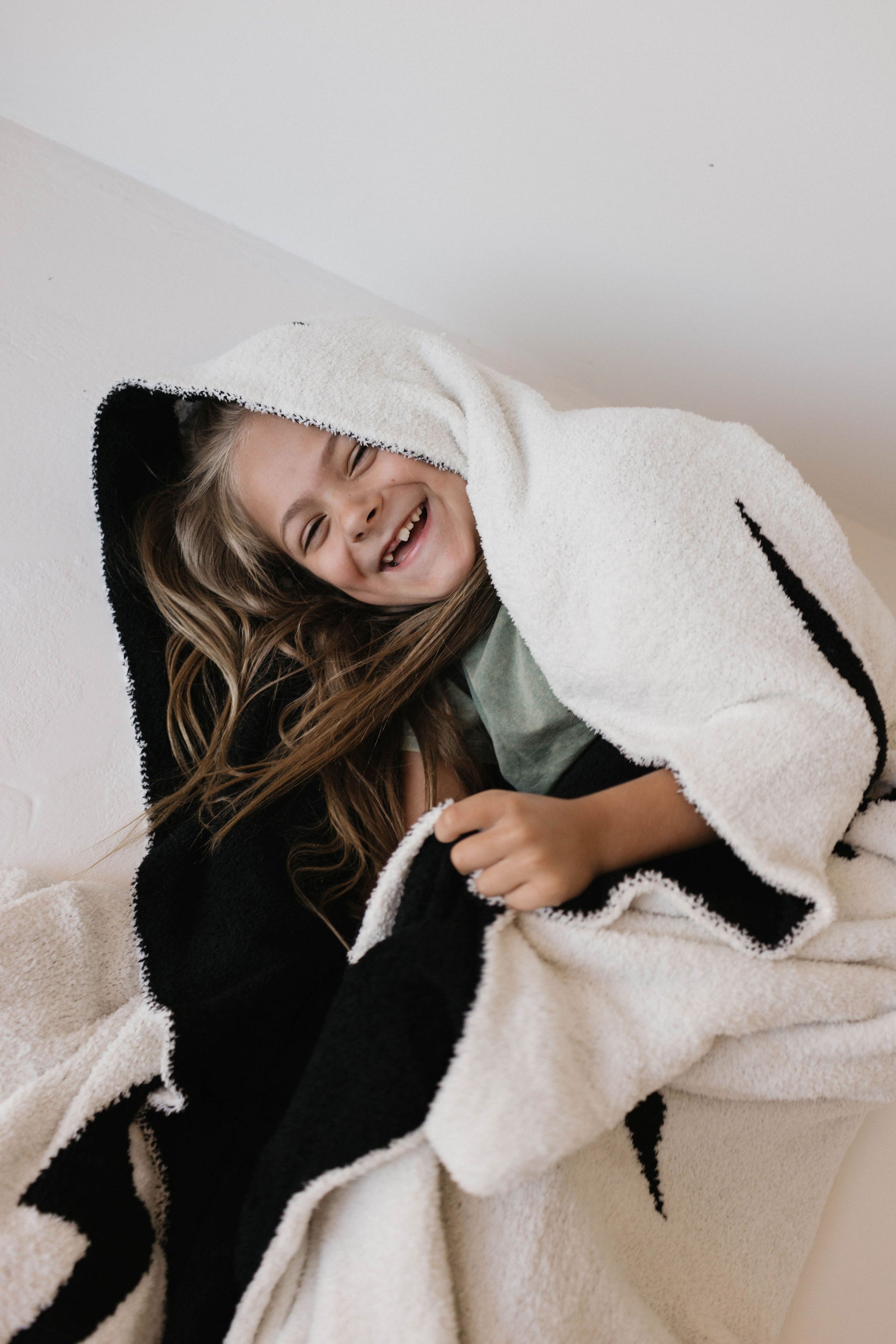 A young girl with long hair is happily wrapped in a dreamy soft blanket. She is smiling broadly and appears to be in a state of joy. The background is plain and light-colored. This 100% Microfiber blanket, the Plush Blanket | Black & White Lightning Bolt by forever french baby, not only offers comfort but is also Oeko-tex certified for your peace of mind.