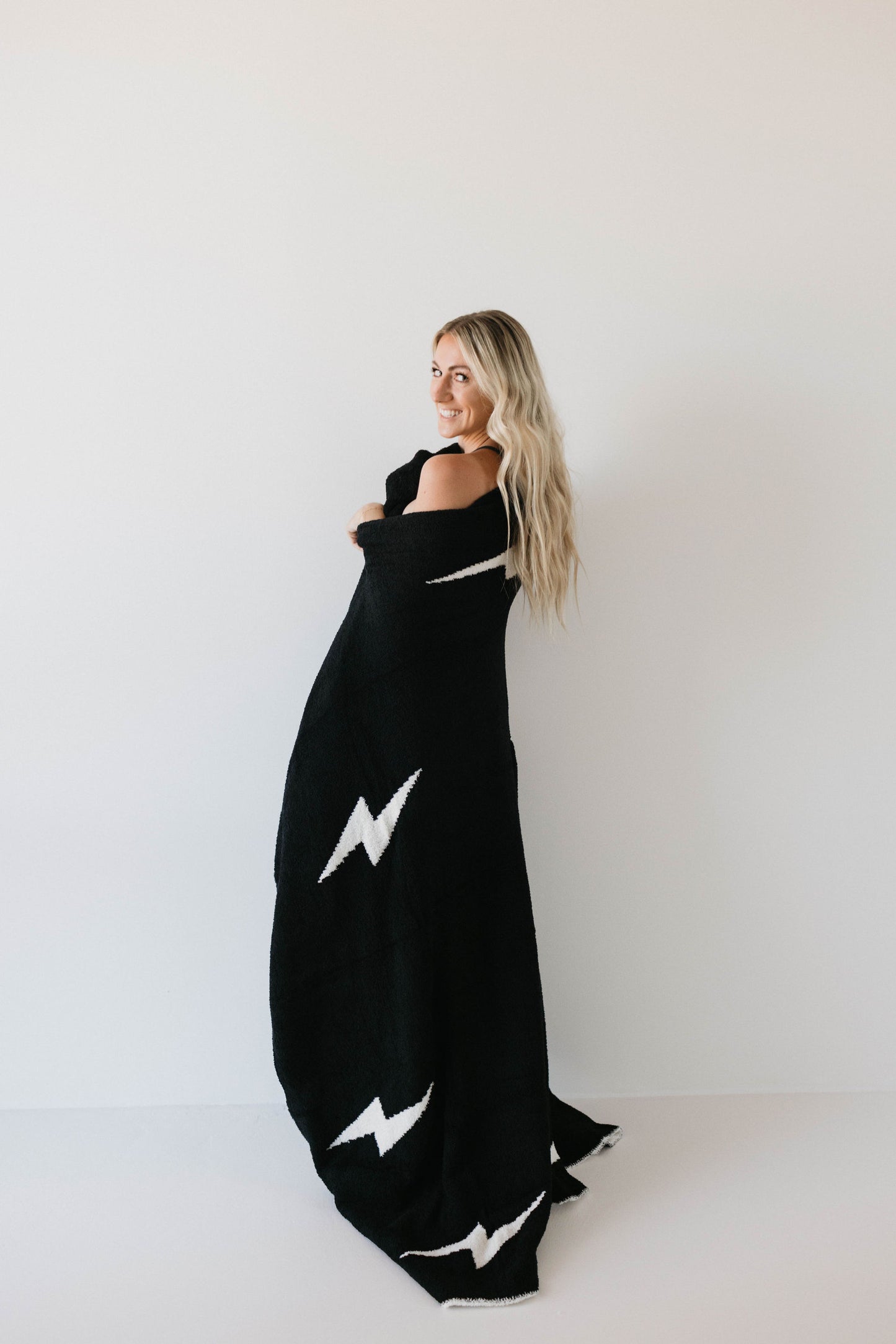 A woman with long blonde hair is wrapped in the dreamy Plush Blanket | Black & White Lightning Bolt from forever french baby, which features striking white lightning bolt designs. She stands sideways, looking over her shoulder and smiling against a plain white background. The black blanket, made from 100% microfiber and Oeko-tex certified for ultimate comfort, enhances the serene scene.