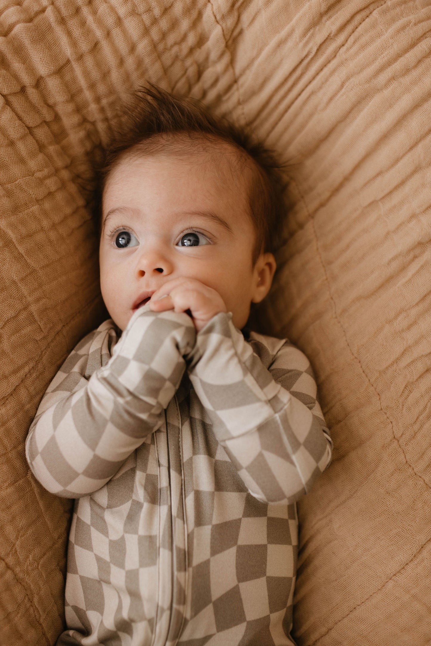 A baby with wide eyes lies on the Latte Quilt from forever french baby. The baby is wearing a beige and white checkered onesie and has their hands near their mouth. They are looking slightly upwards with a curious expression.